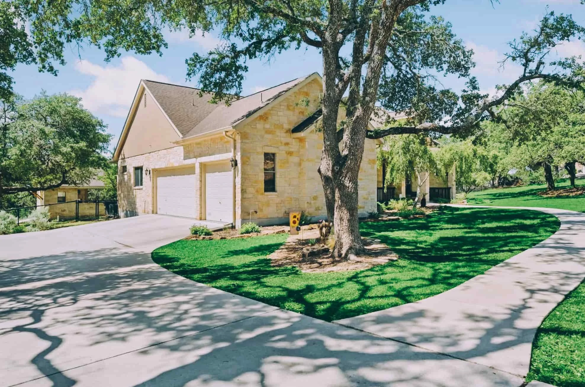 A suburban house with a stone exterior, a two-car garage, a large tree with shade, a green landscaped lawn, and a curved concrete walkway on a sunny day with a partly cloudy sky.