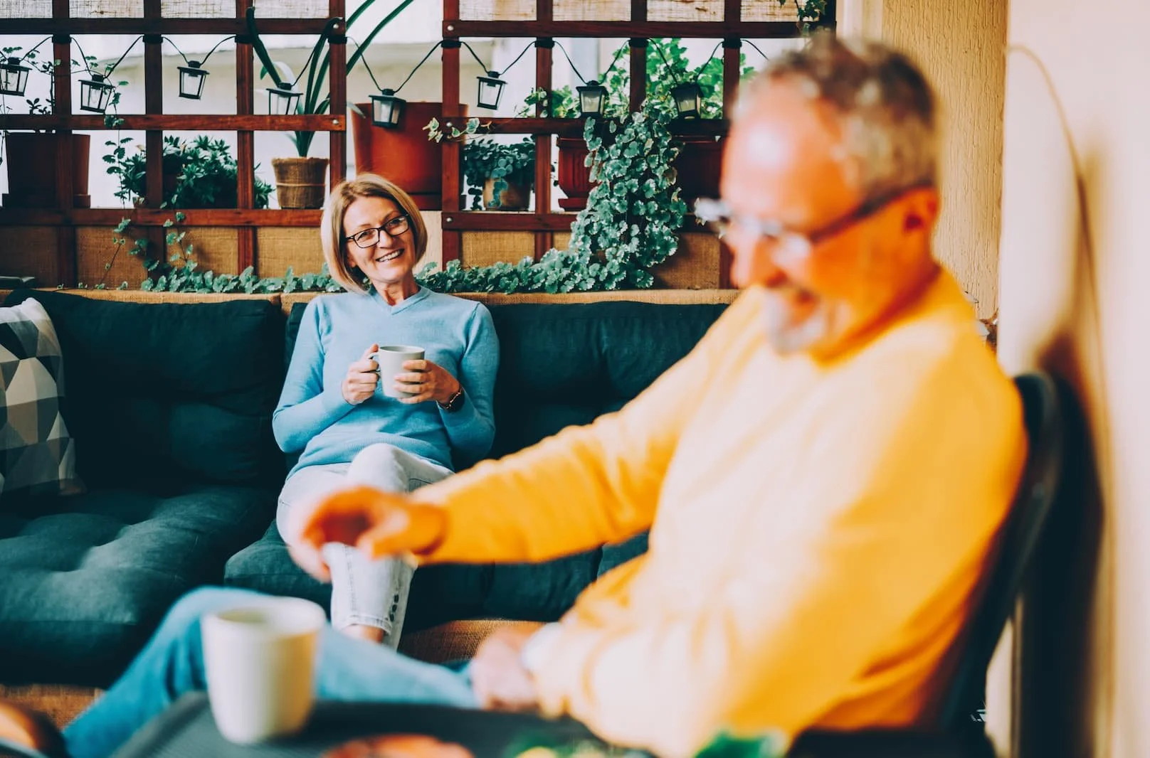 A woman in a blue sweater sitting on a dark green couch, smiling and holding a mug, while a man in a yellow sweater sits beside her with a blurred face, also holding a mug, in a cozy room with plants and decorations in the background.