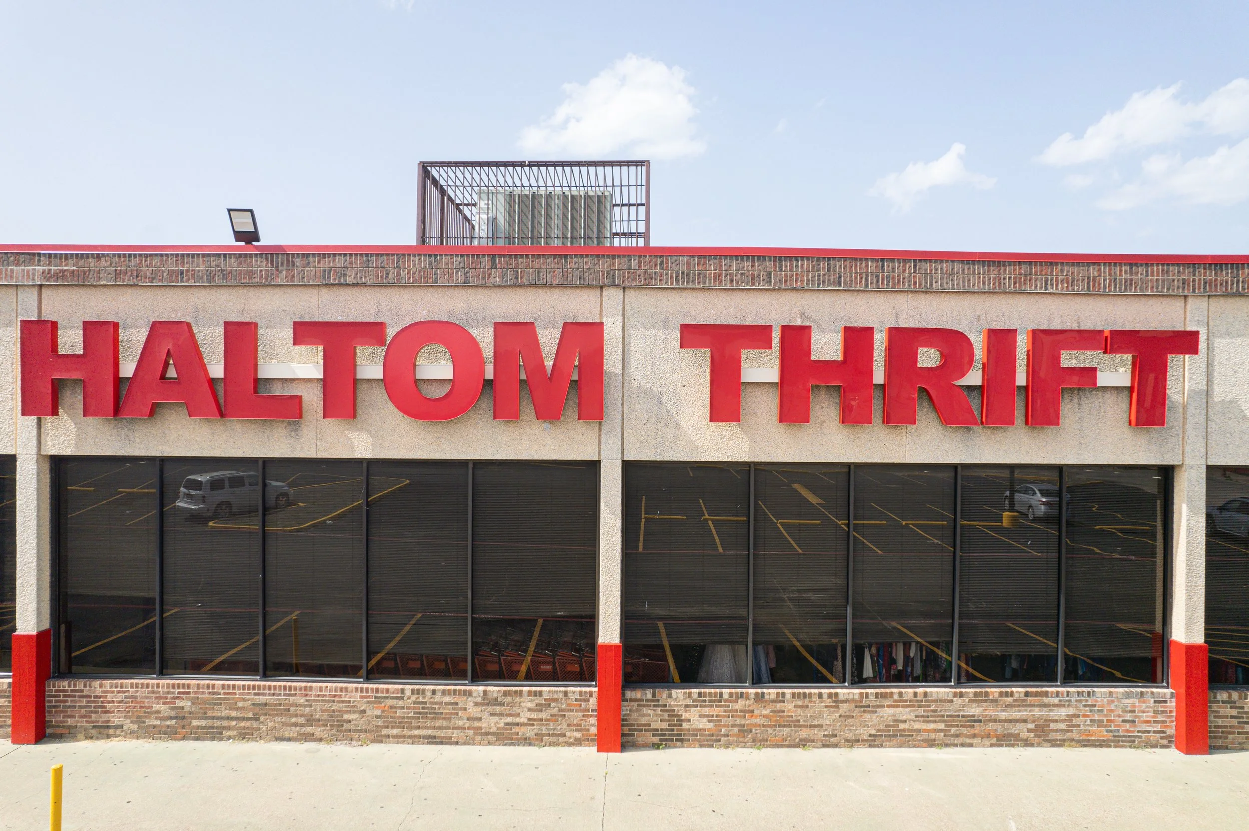 Front view of a retail store with large red letters spelling 'HALTOM THRIFT' on the beige building facade. The building has a row of glass windows reflecting the parking lot and sky, with brick and concrete details.