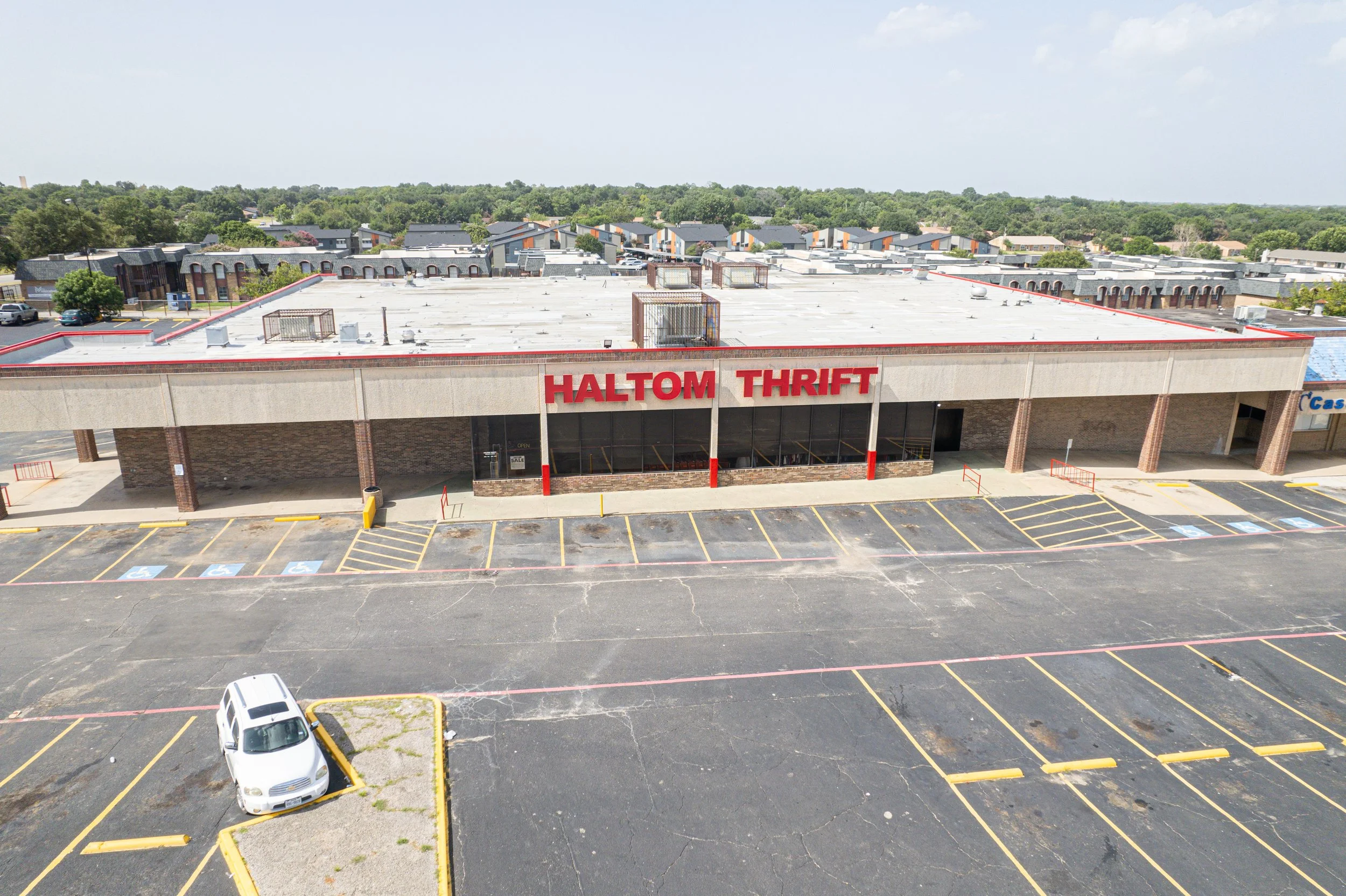 A parking lot and shopping center with a clear blue sky, several parked cars, and visible parking spaces marked with yellow lines. The shopping center has a sign that reads 'HALTOM THRIFT'.