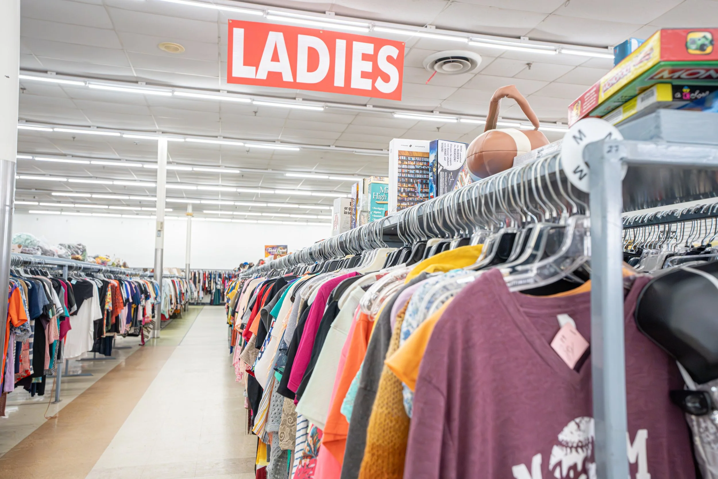 Thrift store aisle with women's clothing under 'Ladies' sign.