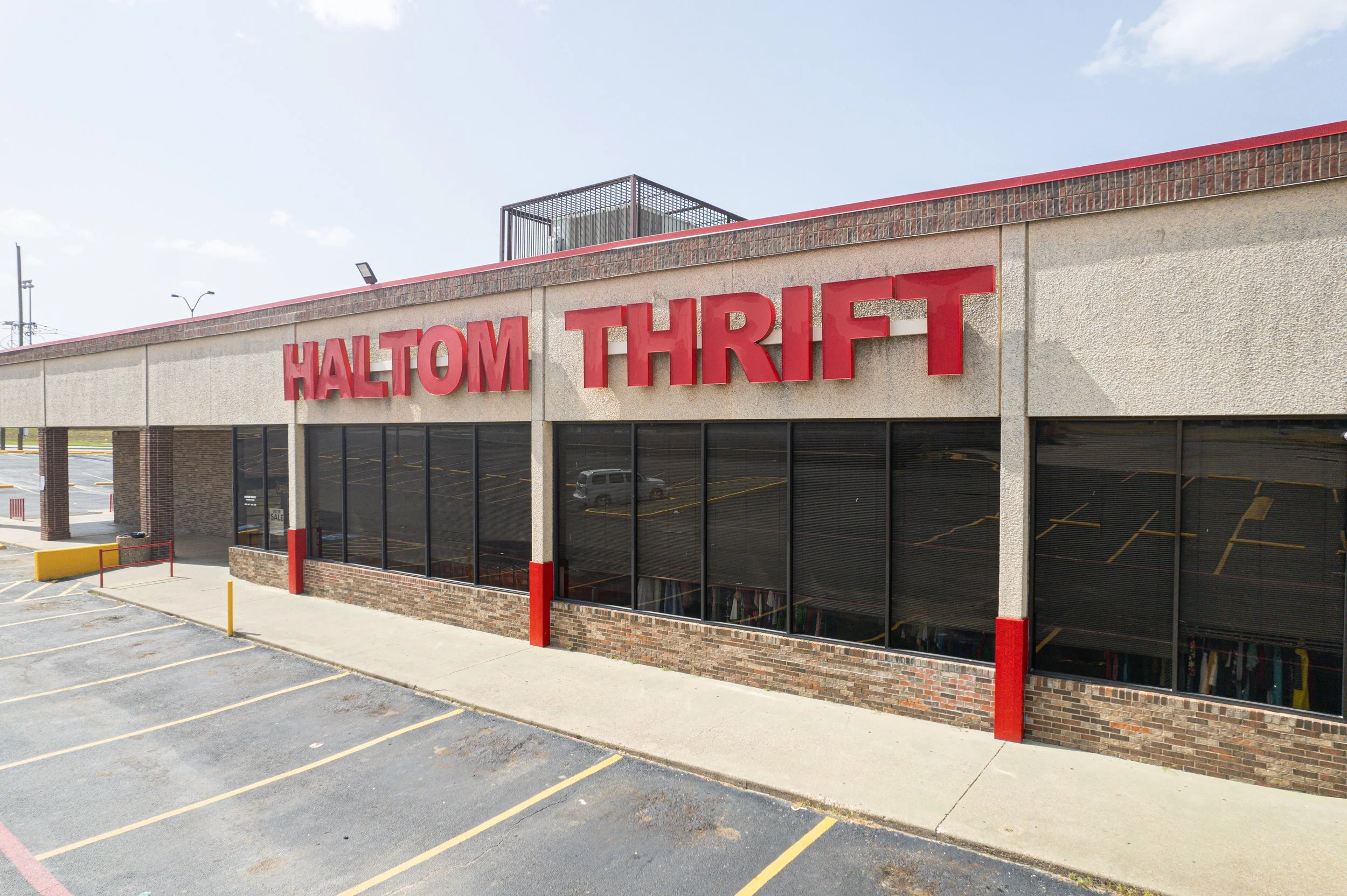 Exterior of a thrift store with large red sign reading 'Haltom Thrift' on a beige and brick building, with an empty parking lot in the foreground and a partly cloudy sky above.