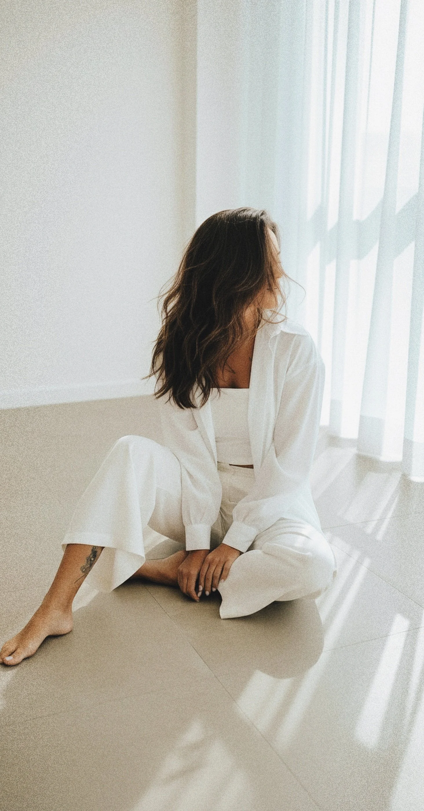 Woman with wavy brown hair in white outfit sitting barefoot on the floor near a window with sheer curtains, sunlight casting shadows.