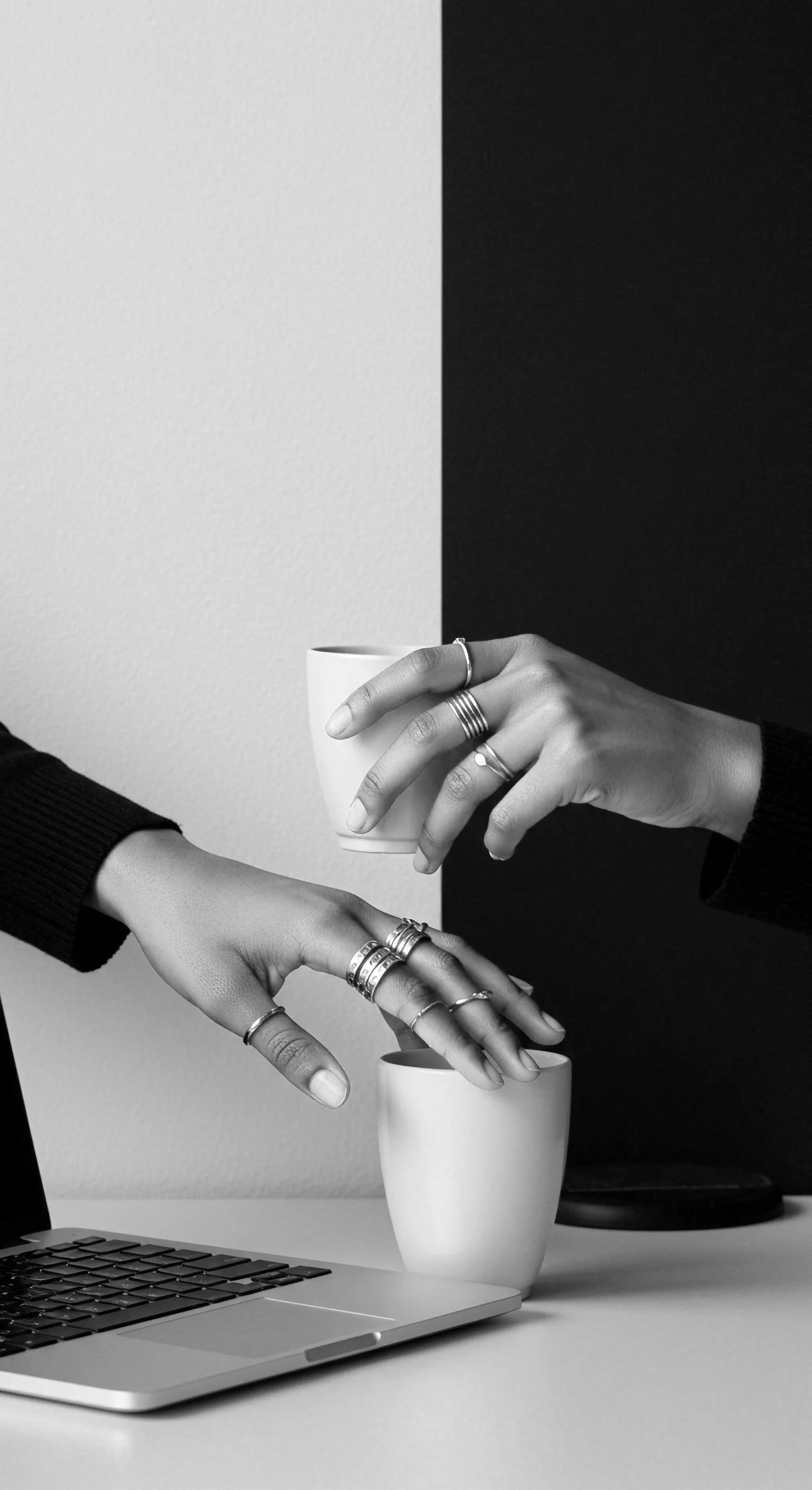 Two hands with rings exchanging a coffee cup in an office setting with a laptop and a second coffee cup on a desk.