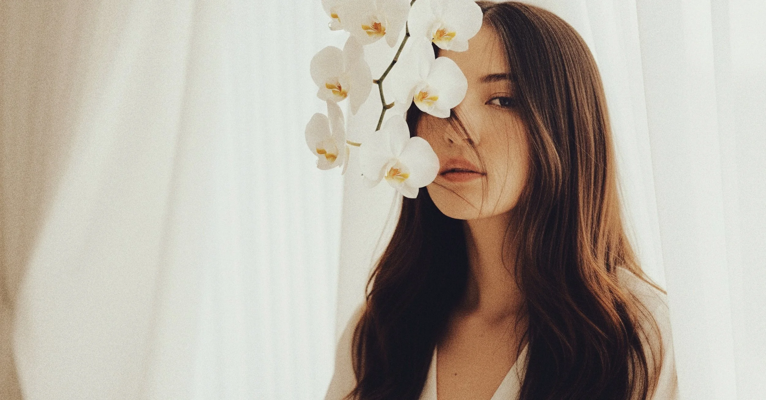 A woman with long brown hair and closed lips, partially obscured by white orchids, standing against a light background.