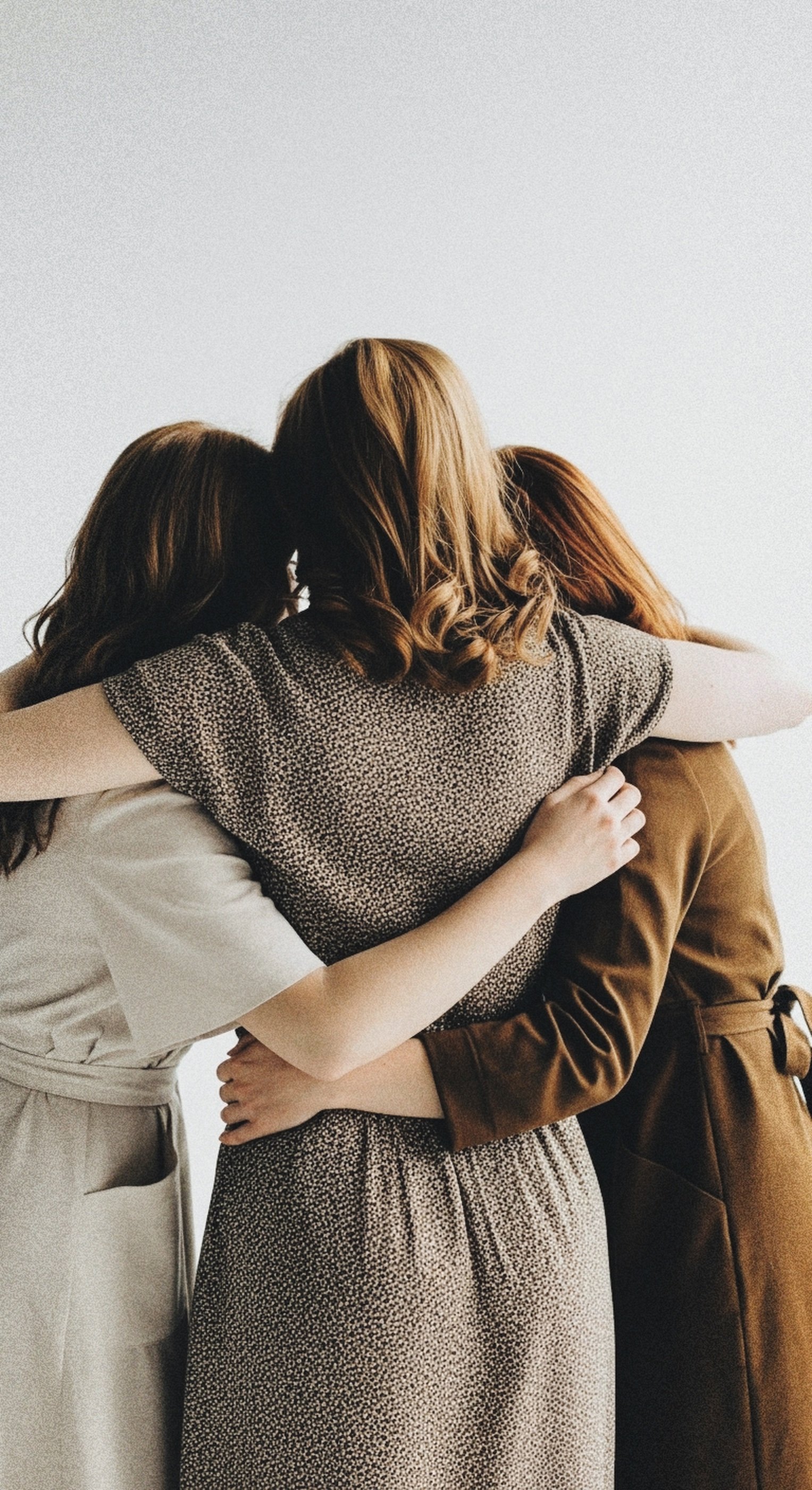 Three women hugging each other against a plain white background, wearing neutral-colored clothing.