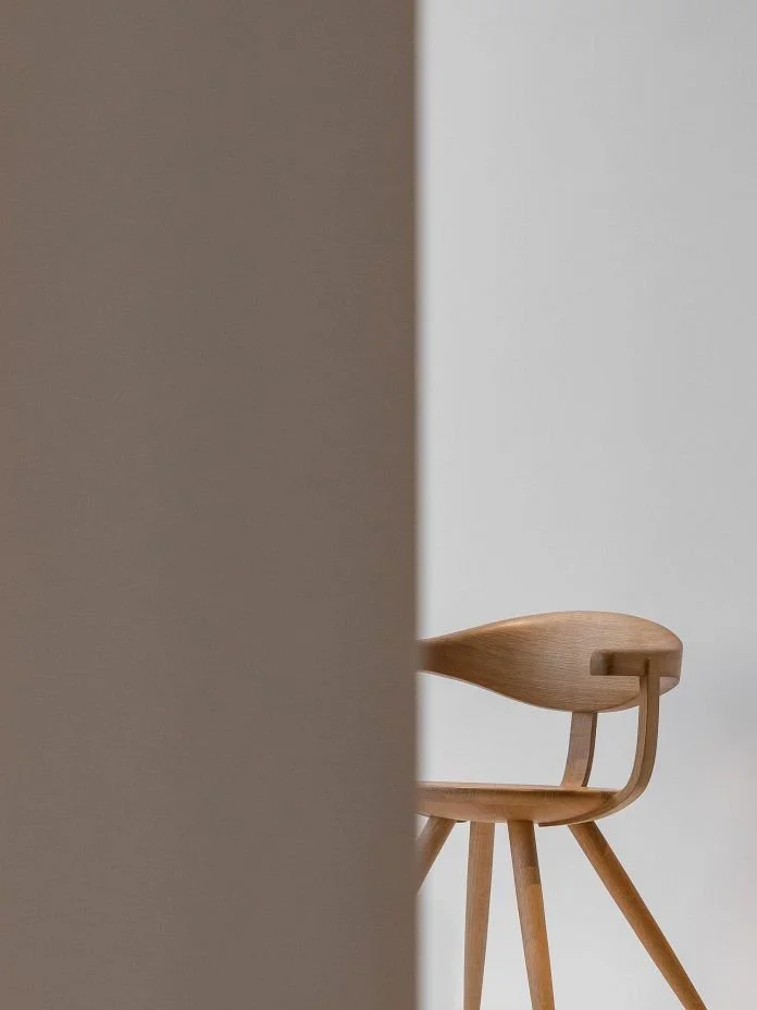 Part of a wooden armchair partially visible behind a beige wall, with a white background.