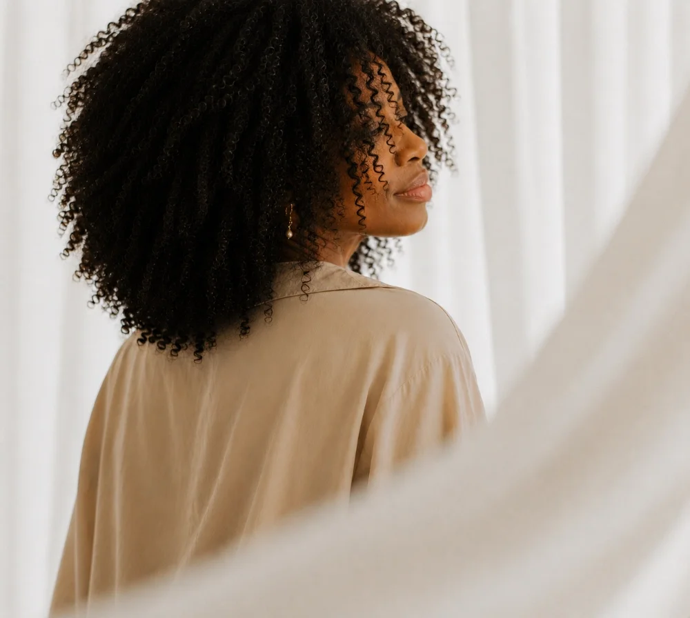 Profile of a woman with curly black hair looking to the side, wearing a beige shirt and earrings, in front of white curtains.