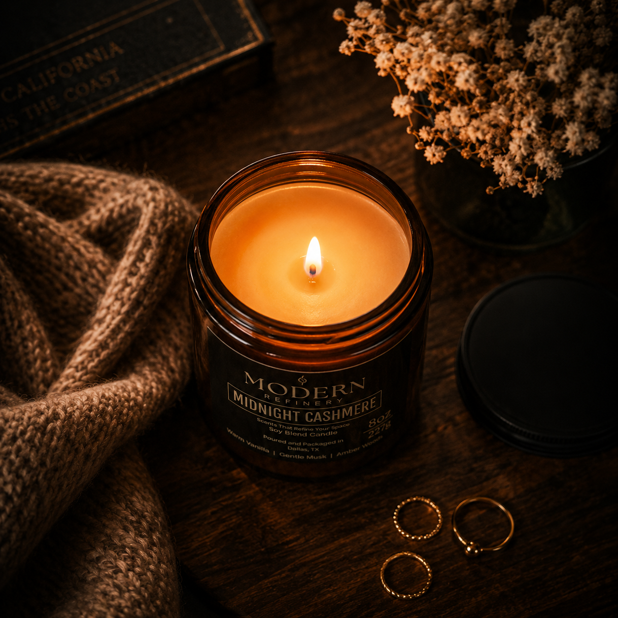 A lit candle labeled 'Modern Refinery Midnight Cashmere' on a wooden surface, accompanied by a knit blanket, a black lid, a small white flower bouquet, and three gold rings.
