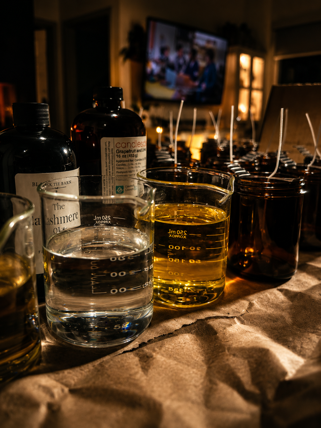 Various candles, including a gingerbread and lemon scent, in glass jars and bottles, with a pitcher of water and a small measuring cup in the foreground on a table. In the background, a television is showing a group of people in a room with warm lighting.