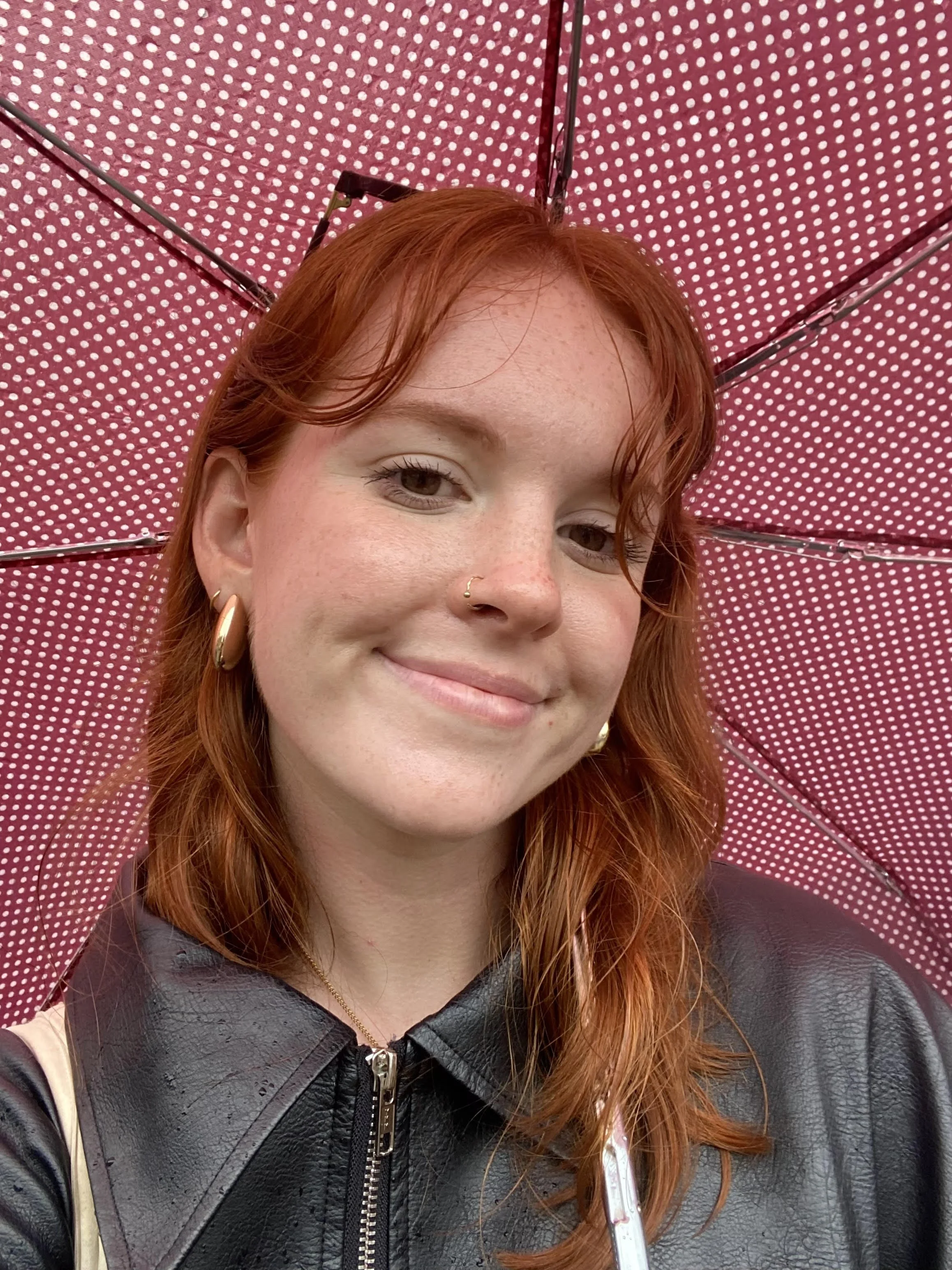 Close-up of a smiling woman with red hair, wearing a black leather jacket, gold hoop earrings, a nose ring, and a gold necklace, holding a red polka dot umbrella.