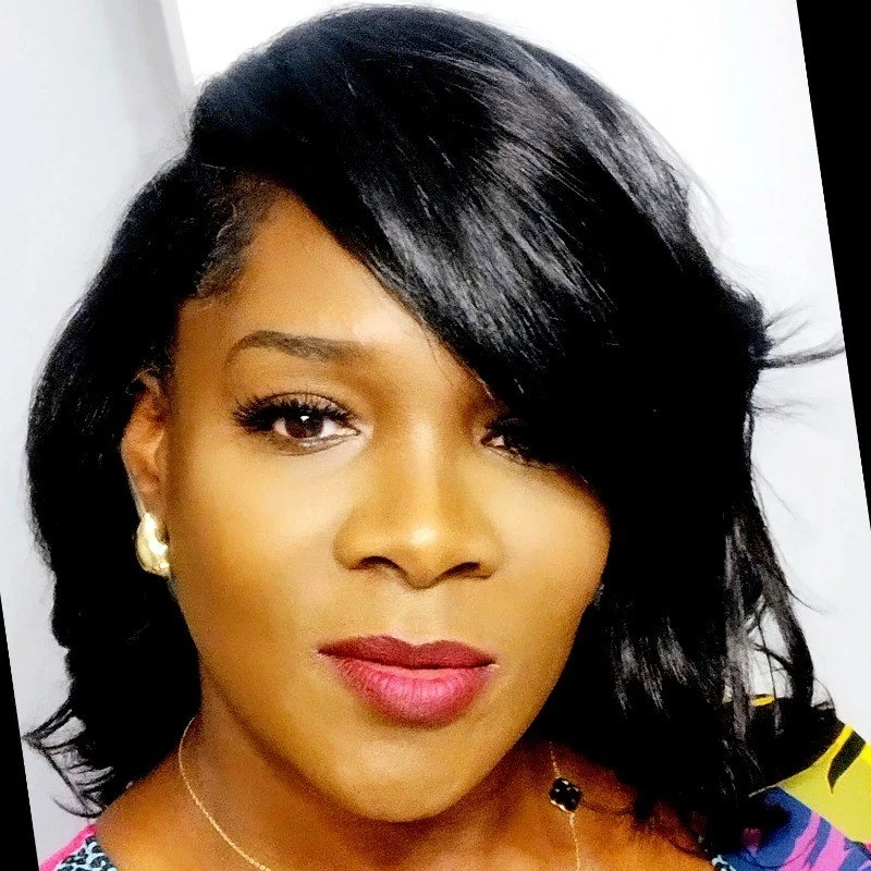 A woman with shoulder-length black hair styled to the side, wearing makeup with red lipstick, gold earrings, and a patterned top, looking directly at the camera.