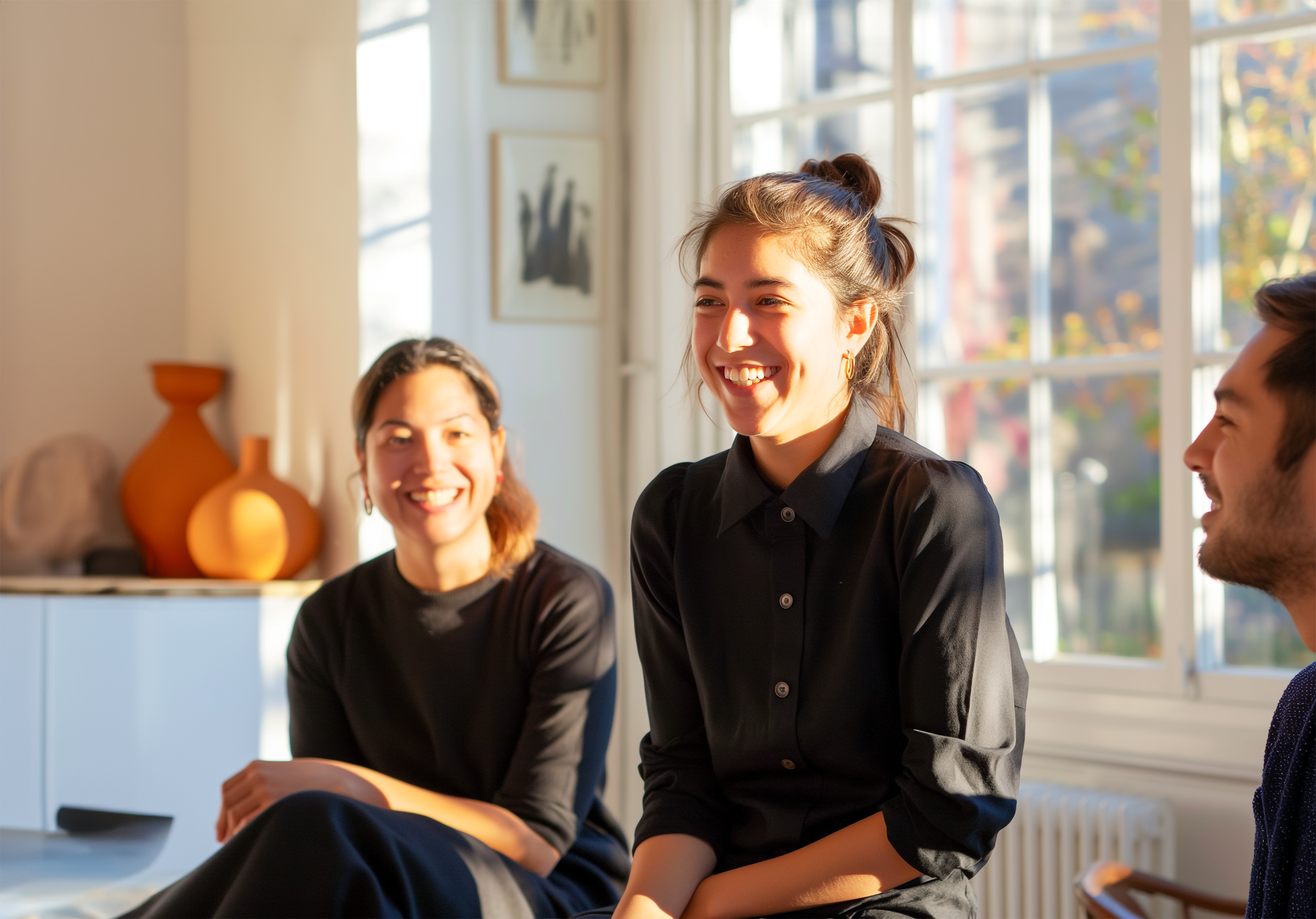 A group of people sitting and smiling in a bright room with large windows, sunlight streaming in.