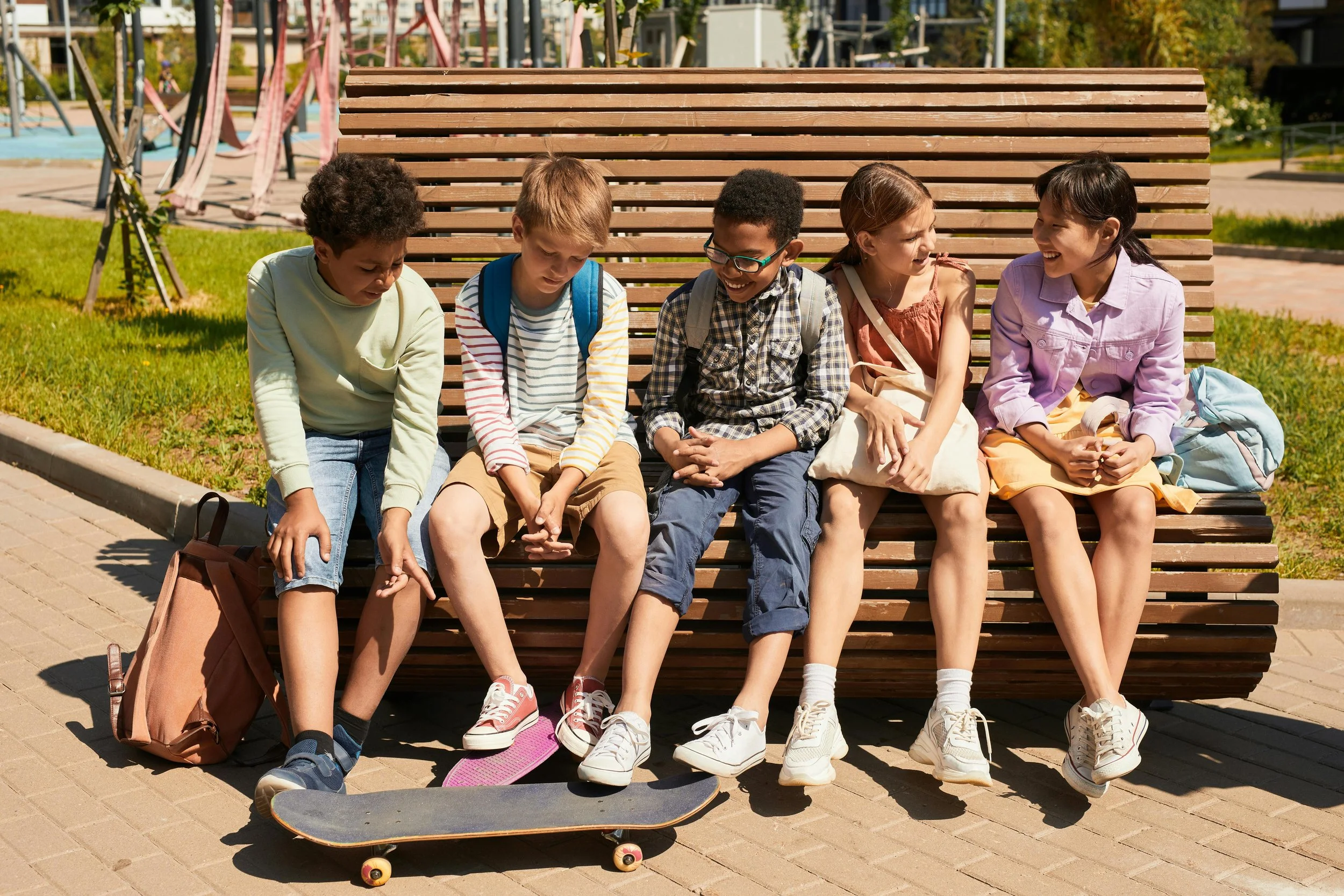 A group of five children sitting on a park bench, two of them with skateboards in front of them, smiling and socializing.