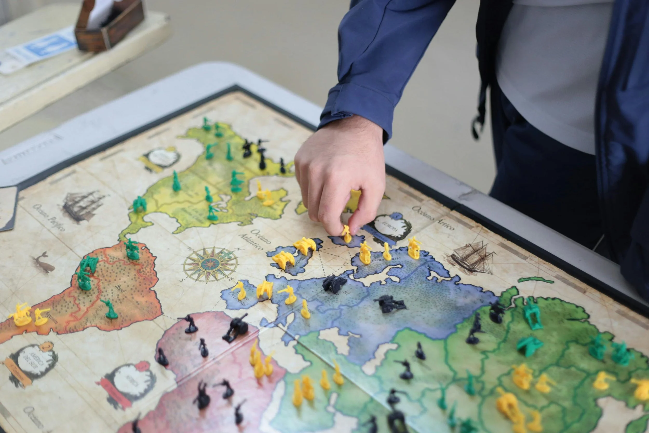 Close-up of a person playing a strategic board game with a world map on the table. The player is moving yellow game pieces and the game includes various colored pieces on the map.