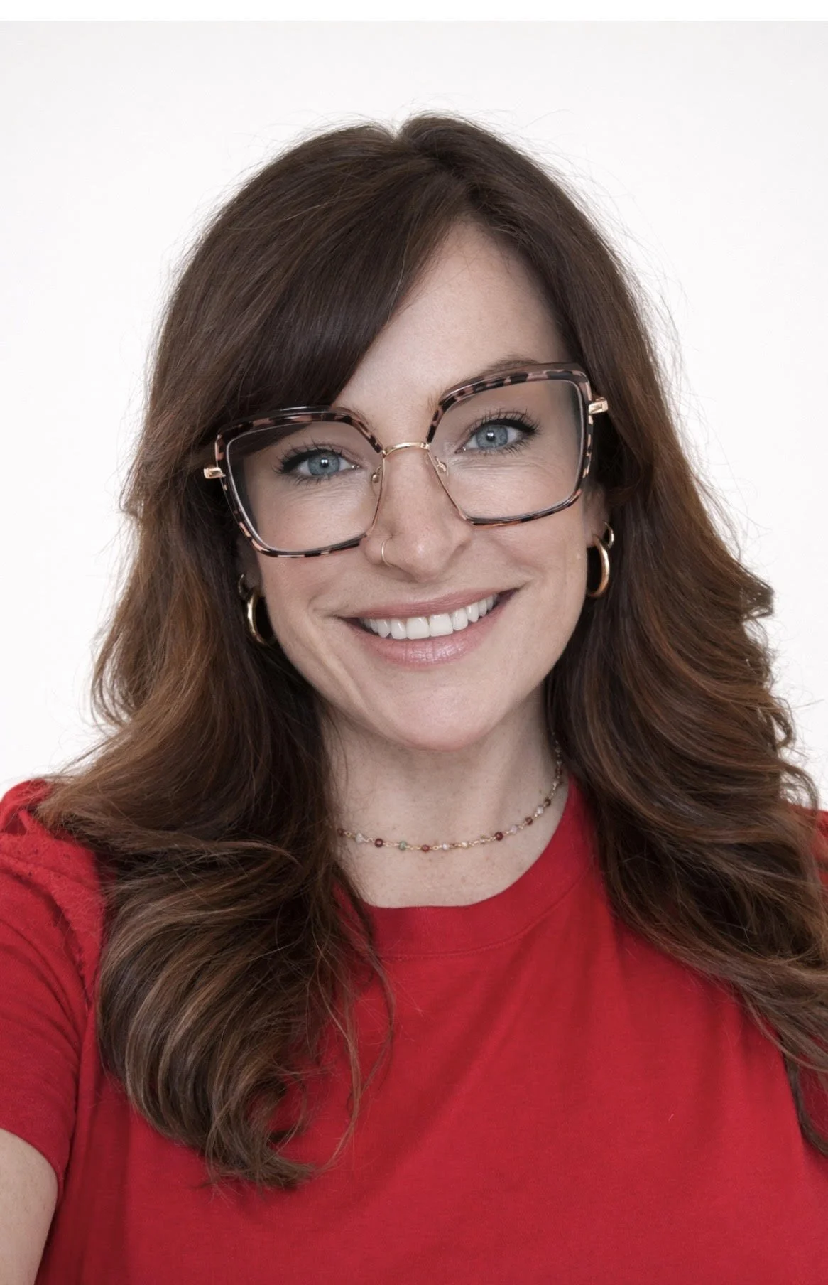 A woman with long wavy brown hair, wearing large tortoiseshell glasses, a red shirt, and jewelry, smiling against a plain white background.