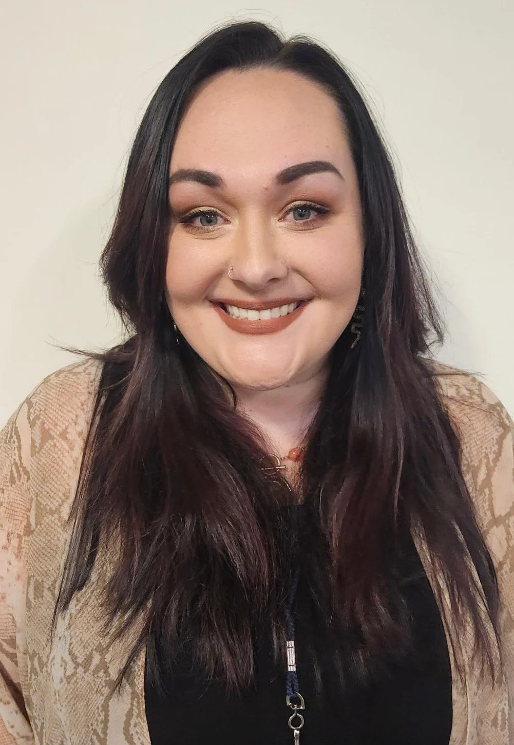 A woman with dark, wavy hair and blue eyes, smiling at the camera, wearing a black top with a lace cardigan and a necklace.
