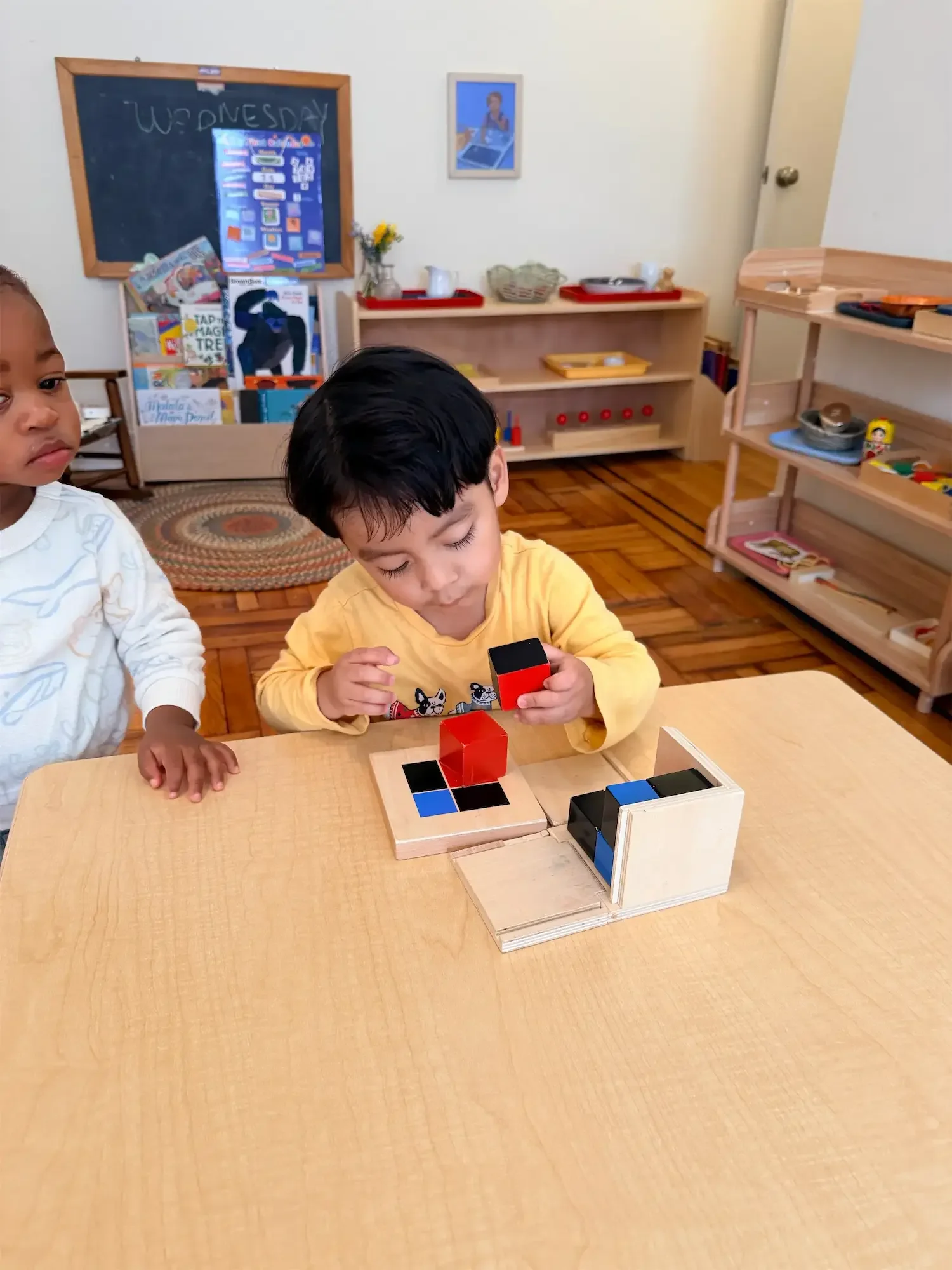 Two young boys sitting at a light wood table in a classroom, playing with colorful wooden blocks. The classroom has wooden shelves with toys and books, a chalkboard with writing, and a framed picture on the wall.