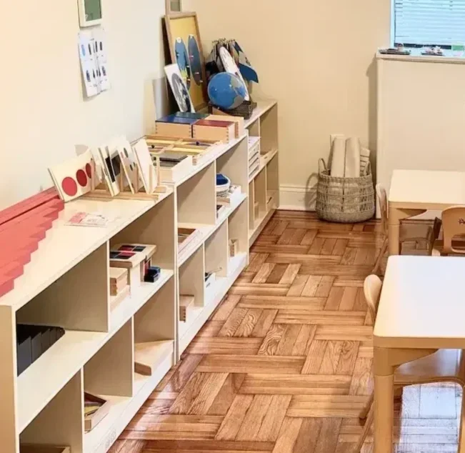 A classroom with white shelves filled with educational materials, books, and artwork, a small table with chairs, and a basket in the corner on a wooden floor.