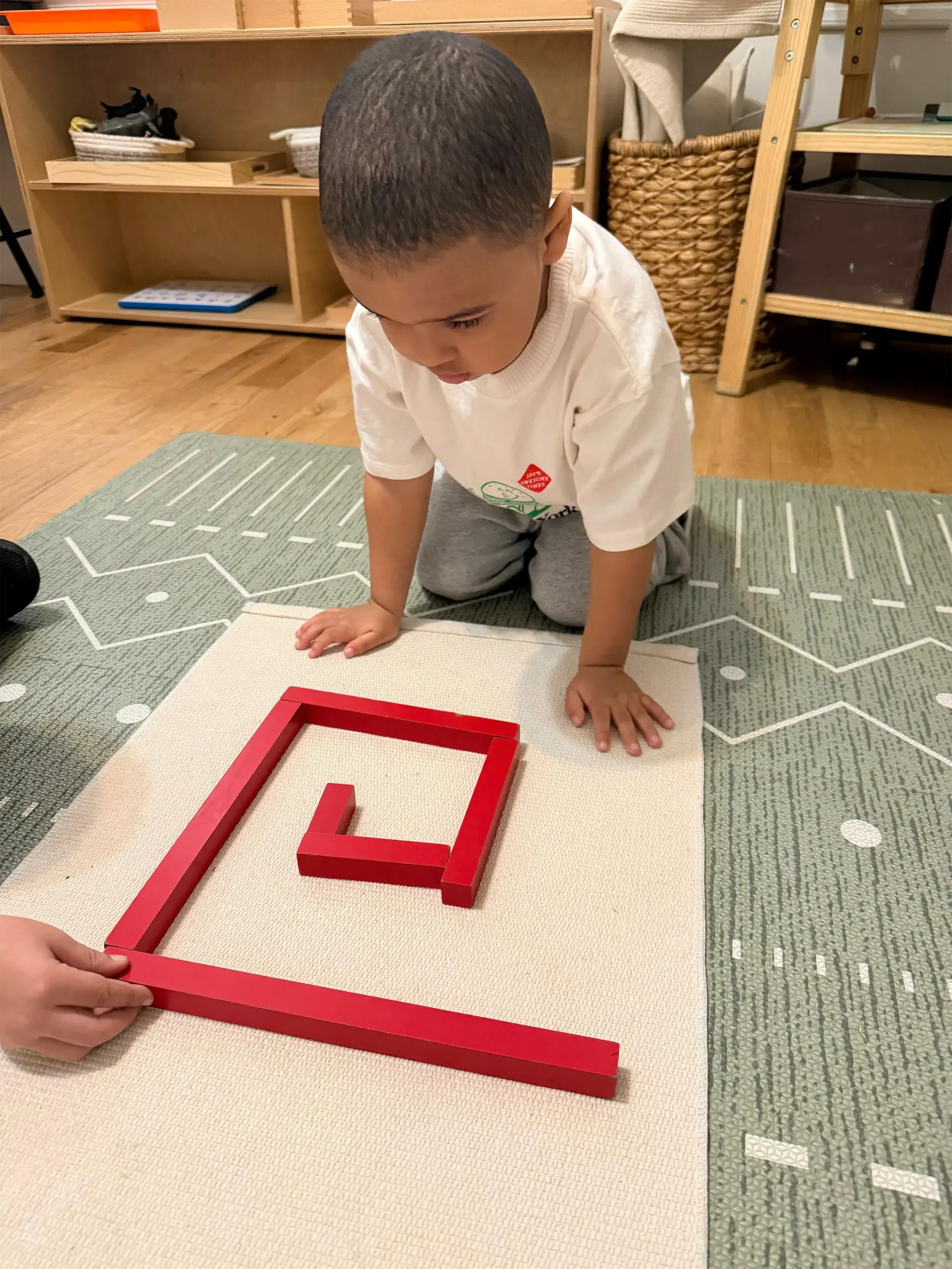 Child in a white t-shirt and gray pants crawling on a patterned rug with a red picture frame on the floor.