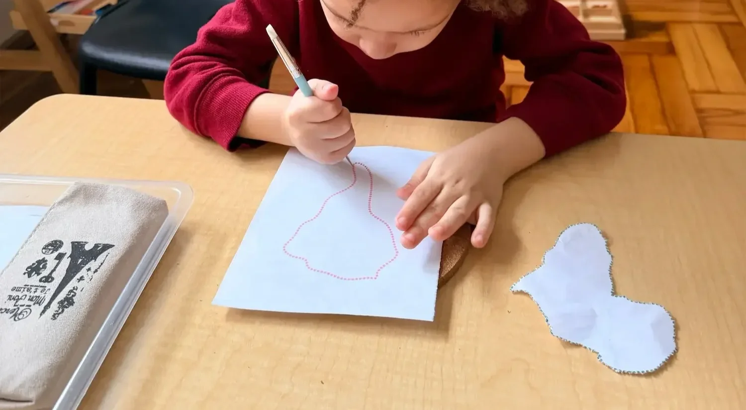 A young child drawing a rabbit outline with a dotted line on a piece of blue paper using a mechanical pencil. There is a paper cutout of the same rabbit shape next to the paper on a wooden table.