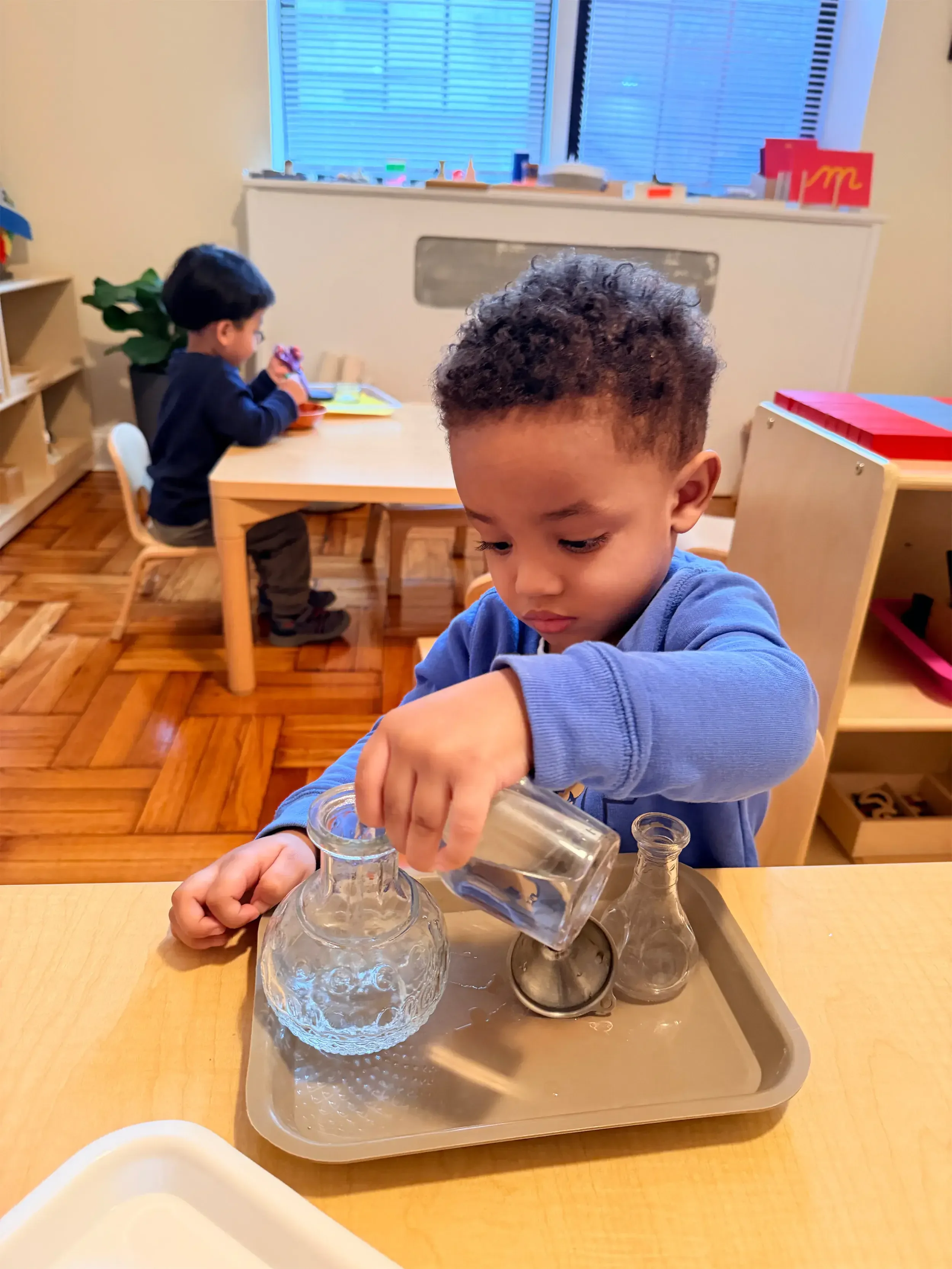 A young boy with curly hair wearing a blue shirt playing with glass laboratory bottles doing practical life montessori work.