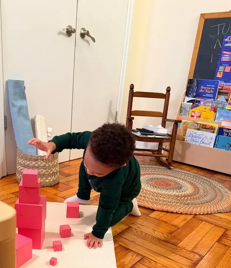 A young child kneeling on a wooden floor, working on the pink tower montessori material.