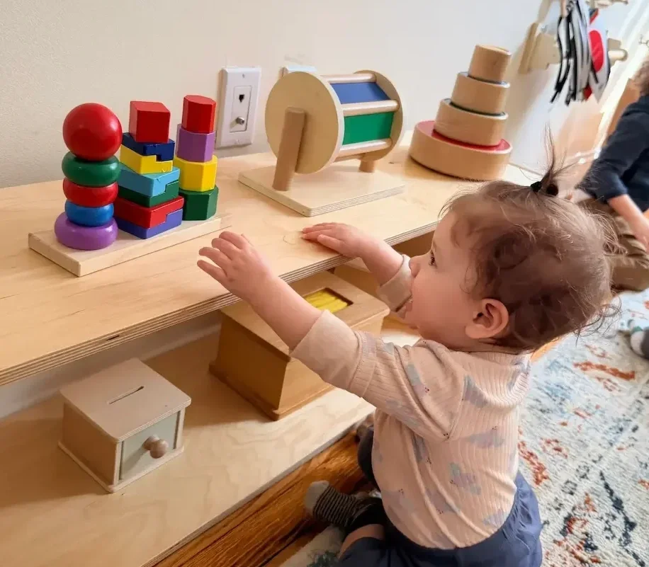 Young child reaching out to colorful wooden stacking toys on a light wood table, in a cozy playroom.