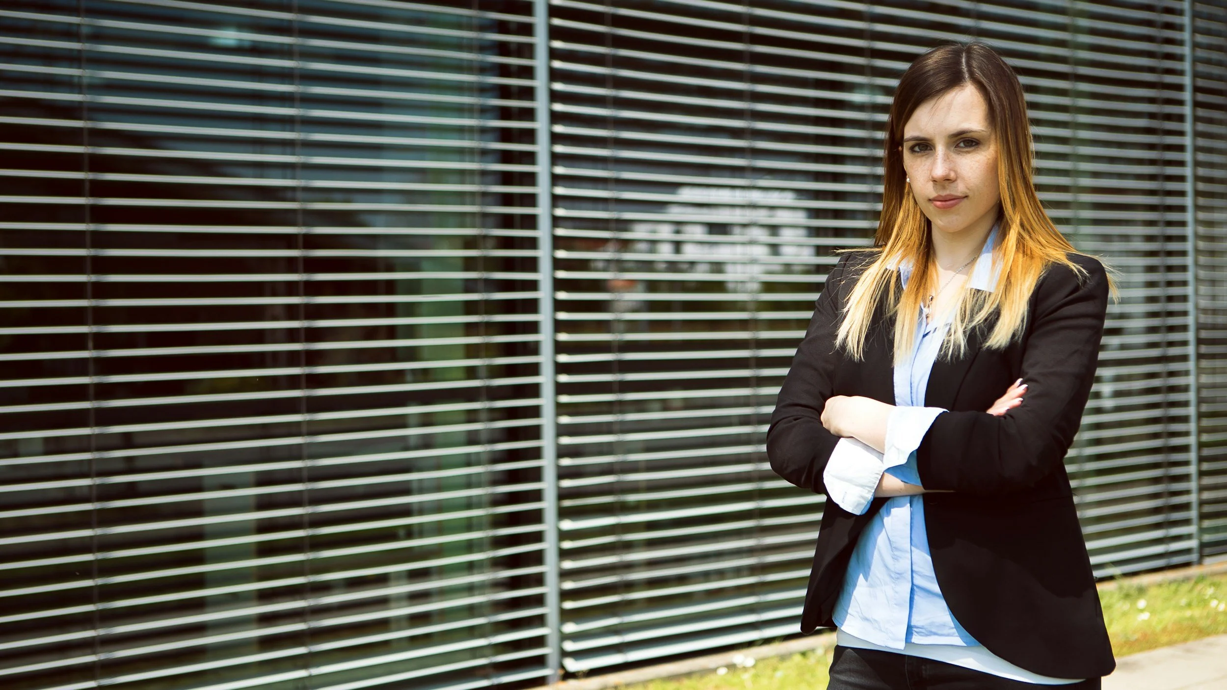 A woman with medium-length brown and blonde hair, wearing a black blazer over a light blue shirt, standing outside with her arms crossed in front of a metal fence.