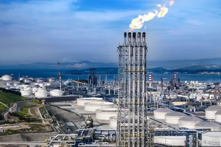 View of an industrial oil refinery with white storage tanks, tall distillation columns, and smokestacks emitting smoke, near a body of water under a partly cloudy sky.