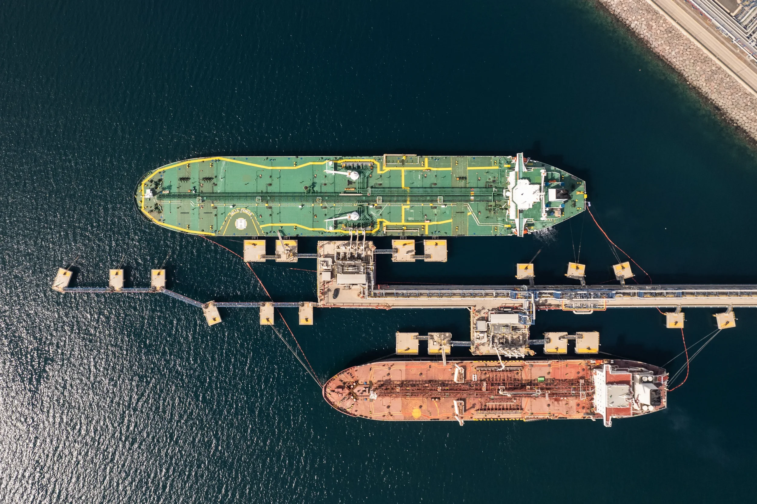 An aerial view of a large cargo ship docked at a port, with water on both sides and a breakwater in the background.