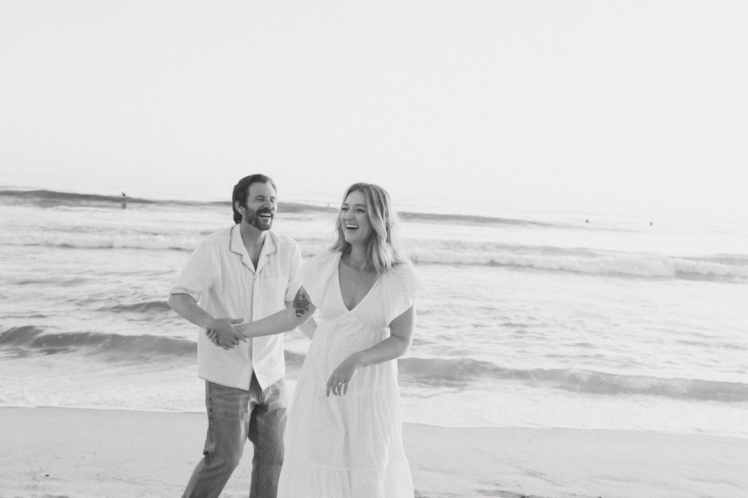 A black and white photo of a couple holding hands and smiling, walking on the beach with ocean waves in the background.