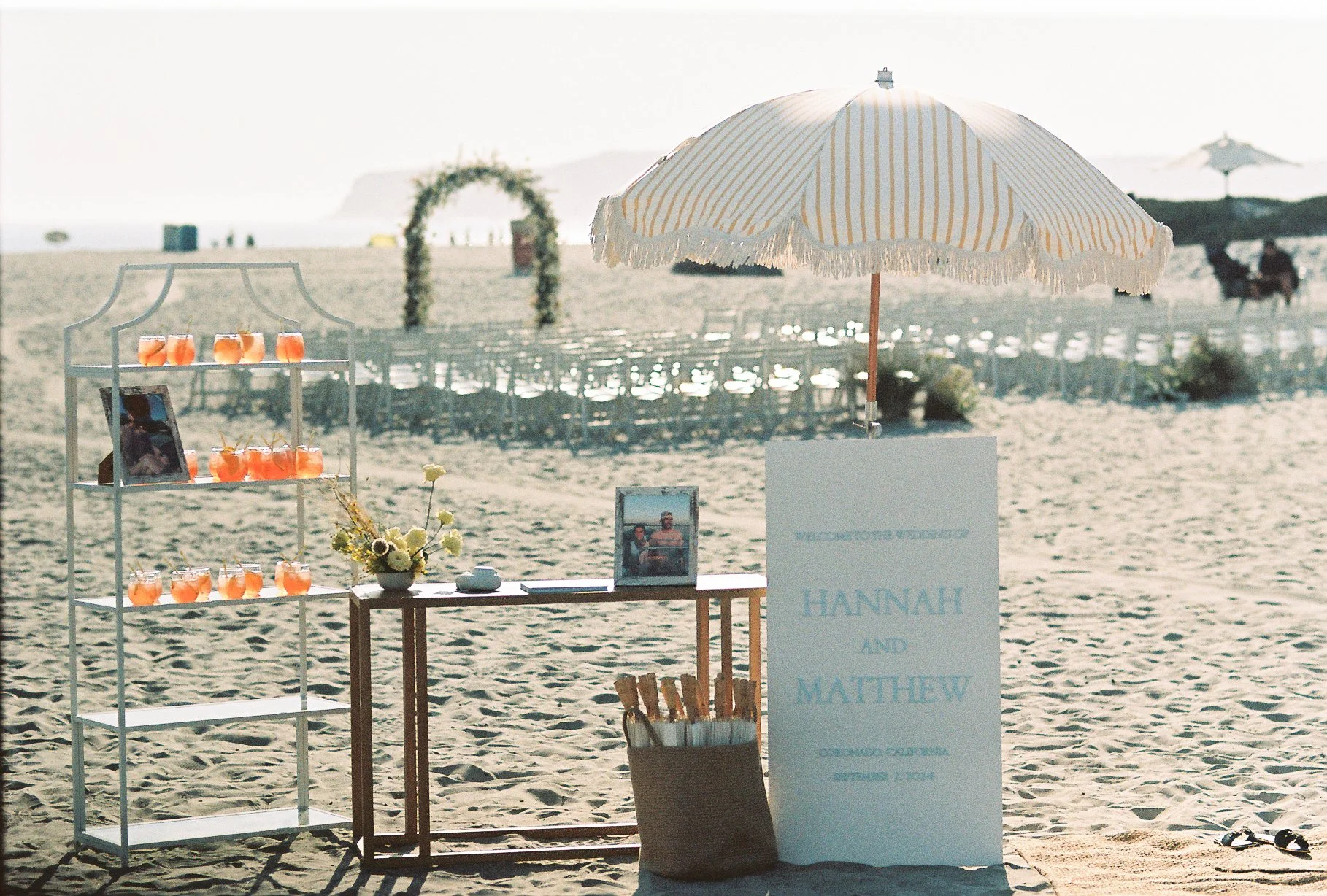 Decorative wedding setup on a beach with a white and yellow striped umbrella, a display shelf with orange-colored drinks, a framed photo, a bouquet of flowers, a sign with the names Hannah and Matthew, and chairs in the background under beach umbrell