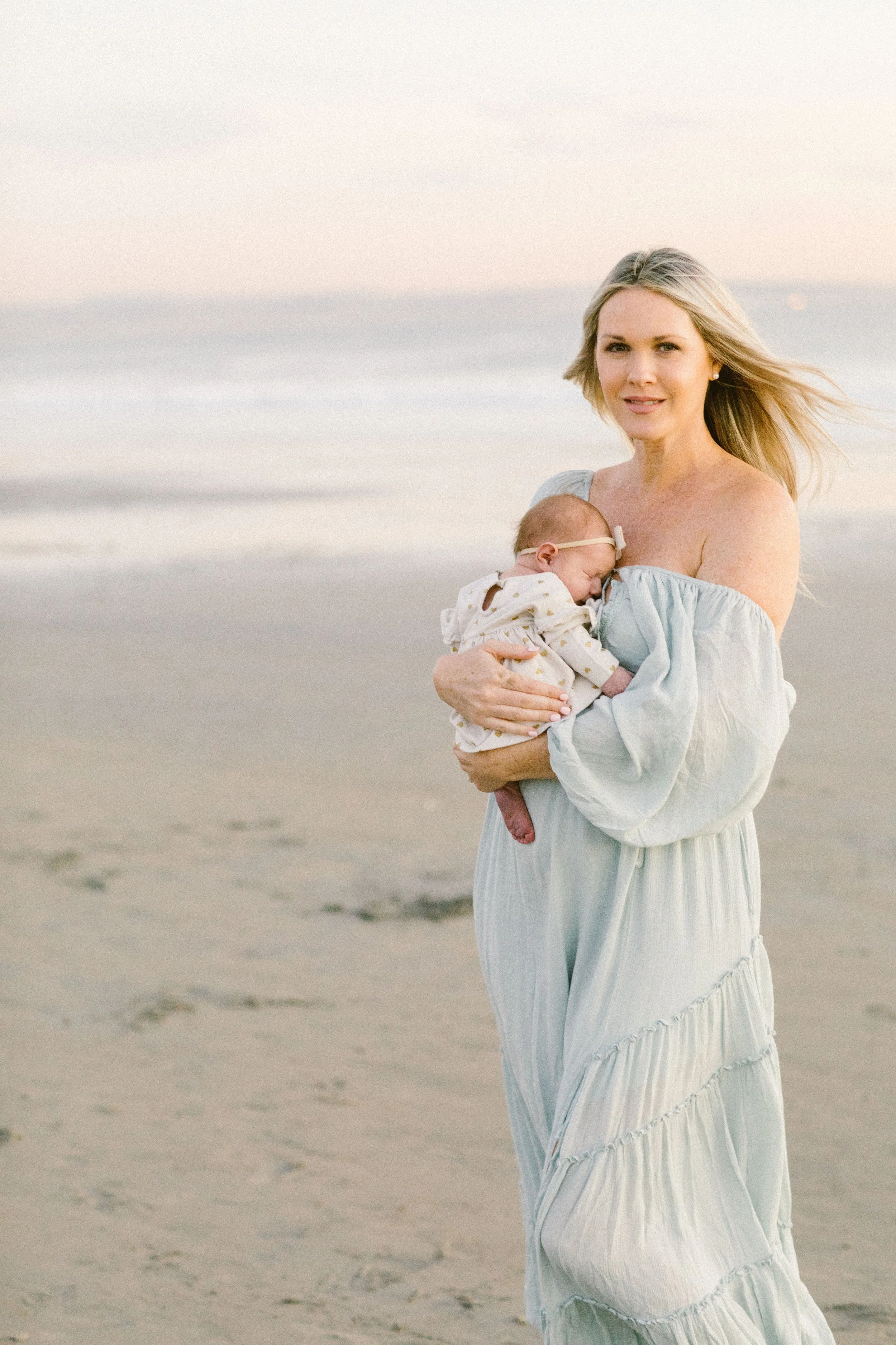 A woman in a light blue dress holding a newborn baby on a beach with the ocean in the background.