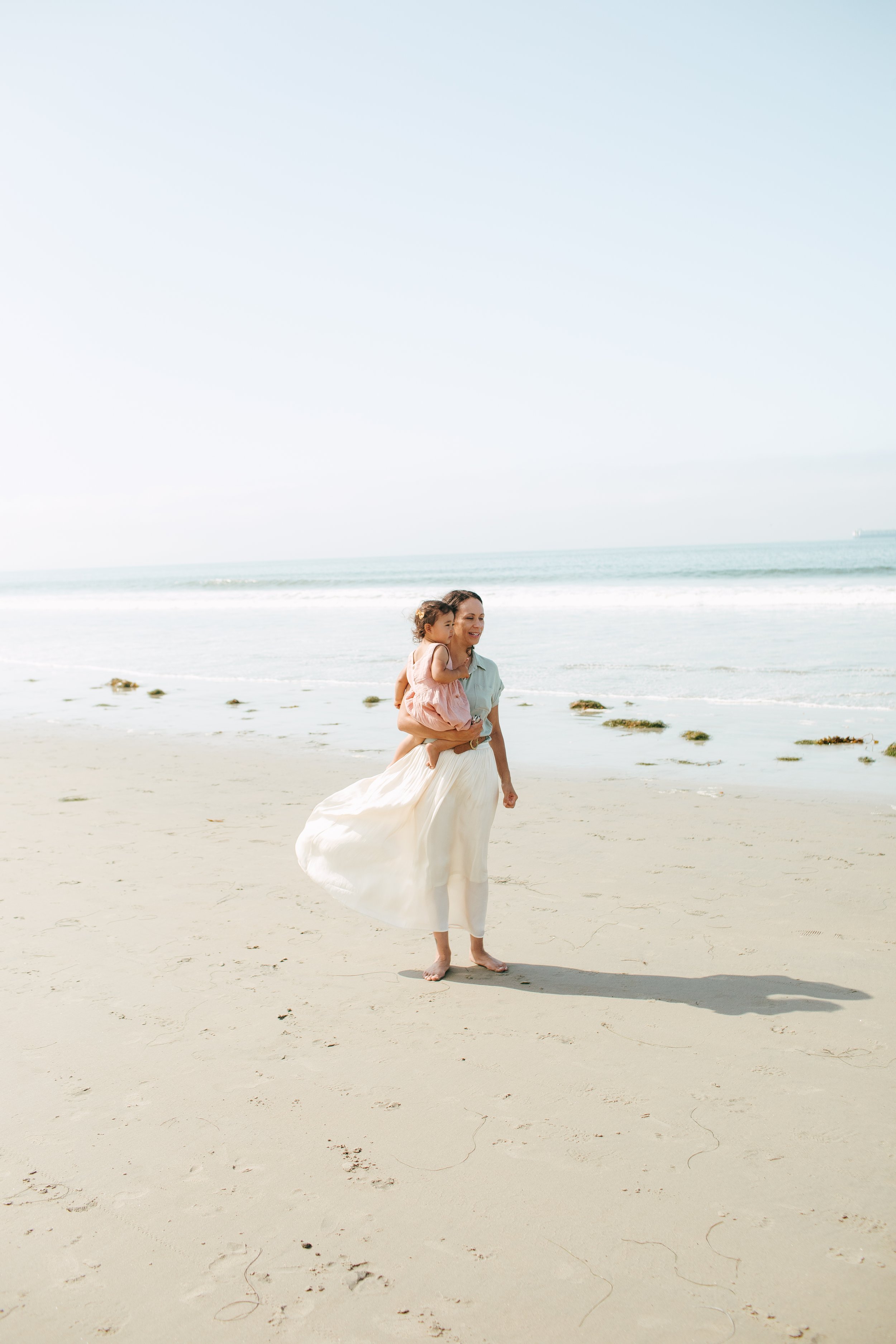 A woman carrying a young girl on her shoulder walking on a sandy beach near the ocean with a clear sky in the background.