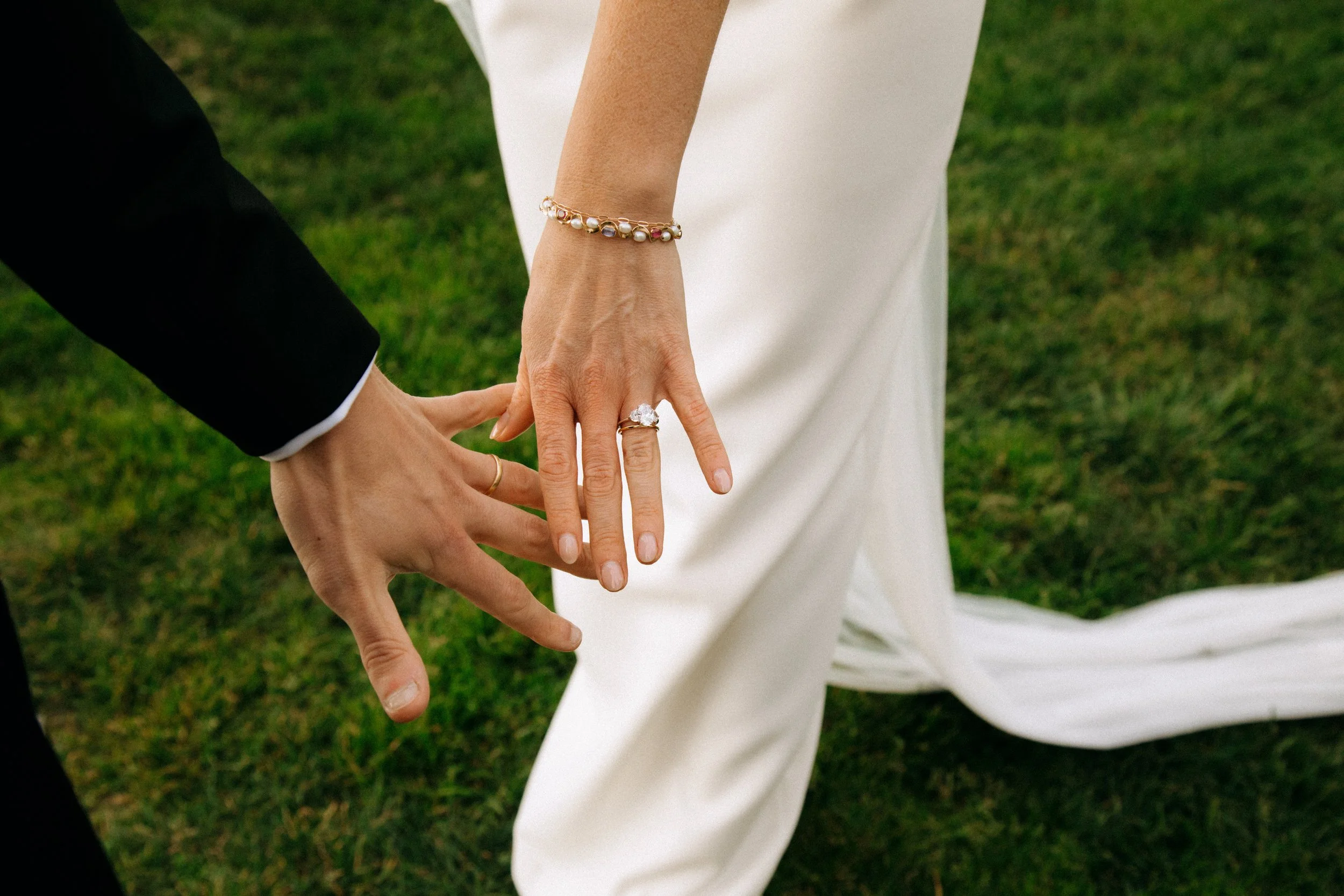 Close-up of a person wearing a pearl bracelet and a large ring, holding hands with another person in a black suit, outdoors on green grass.