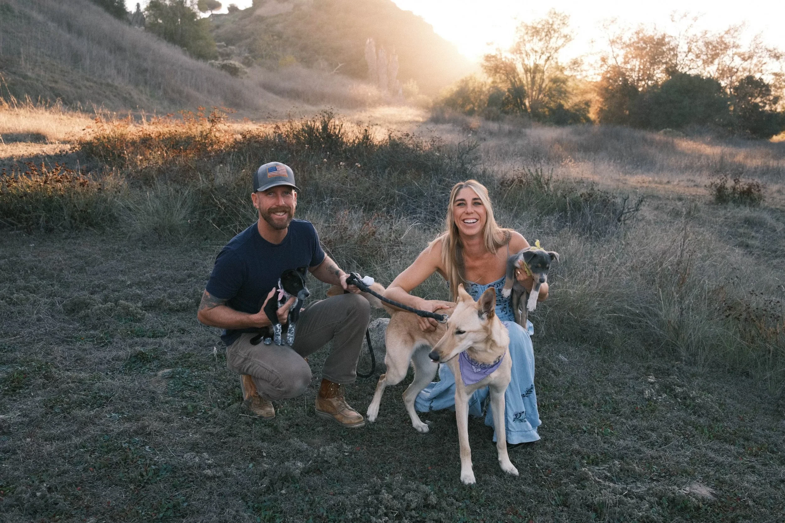 A man and a woman smiling while holding dogs in a grassy, outdoor area during sunset with trees and hills in the background.