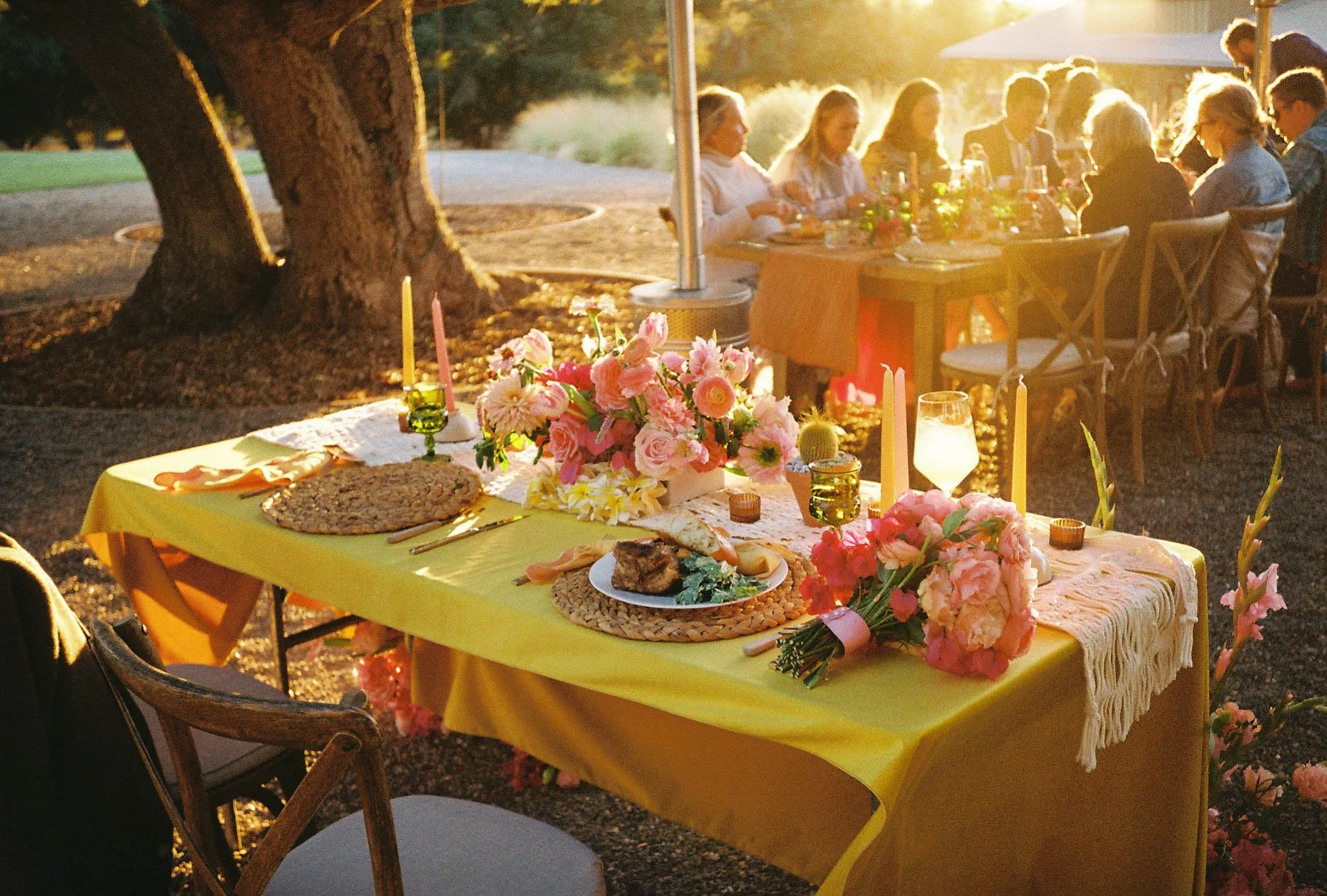 Dinner table outdoors during sunset with pink and white flowers, candles, and food, surrounded by people sitting and enjoying the meal.