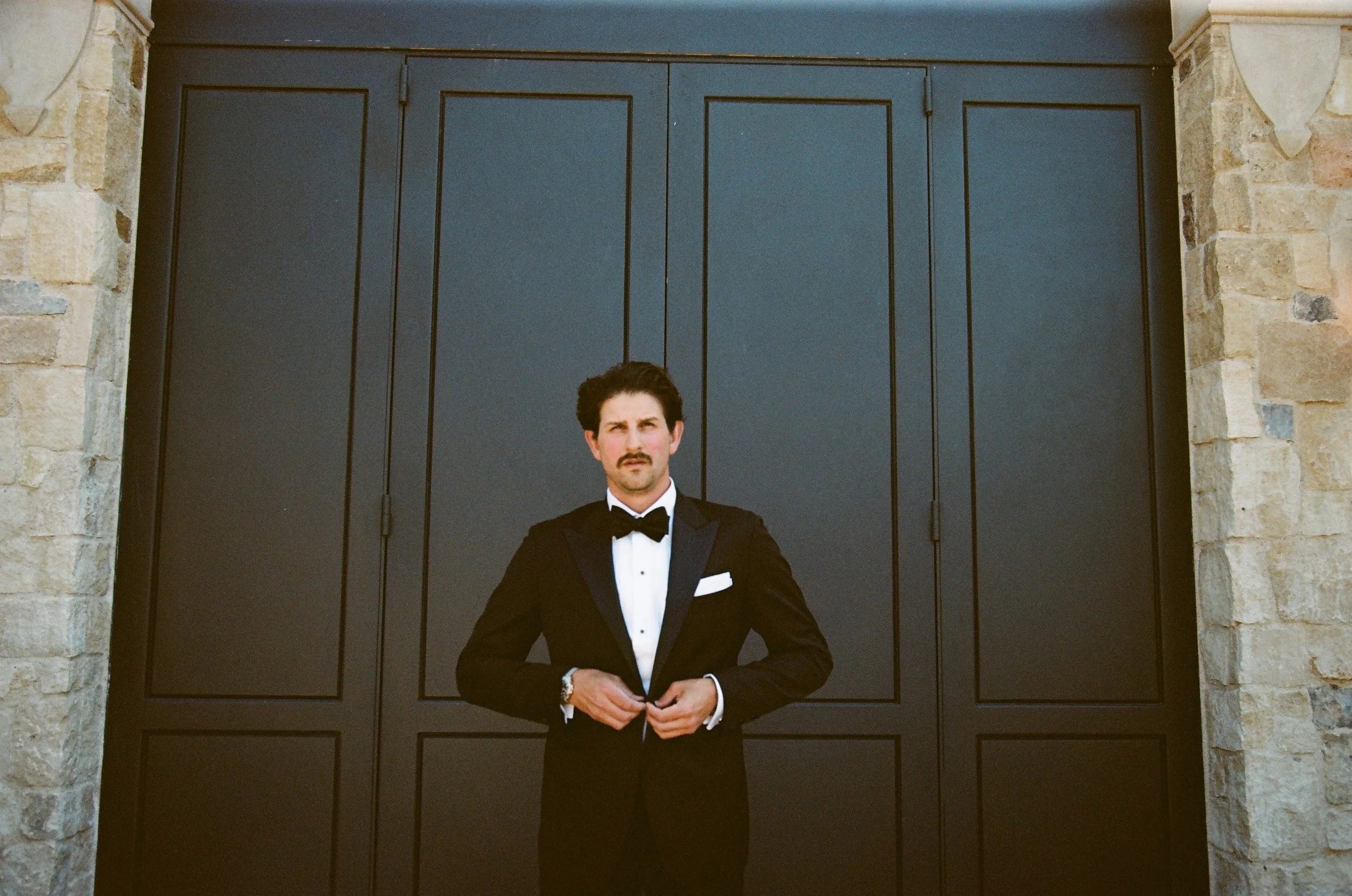 A man in a black tuxedo with a bow tie standing in front of a large dark gray door framed by stone walls.