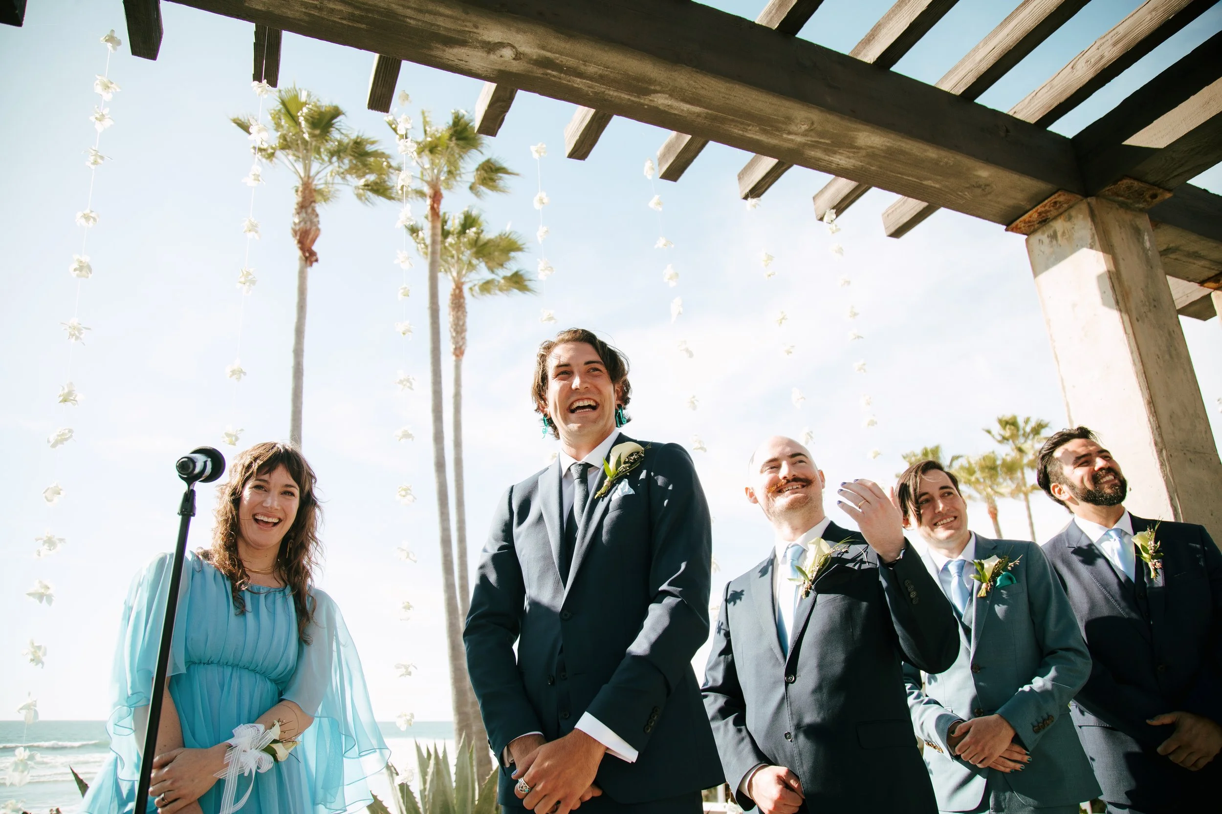 A group of people at a wedding ceremony outdoors, under a wooden pergola decorated with hanging white flowers. Palm trees and a blue sky are visible in the background.