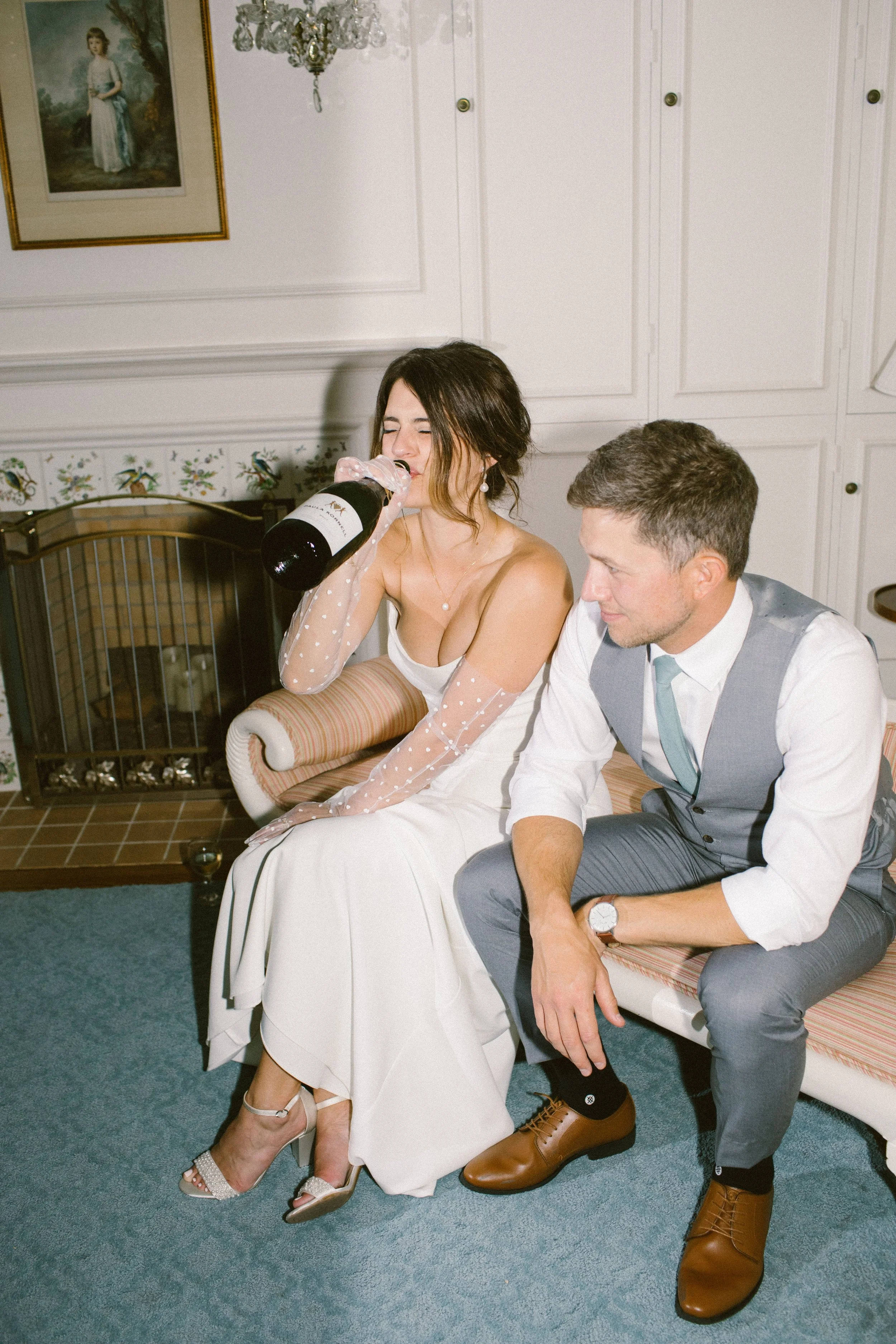 A woman in a white dress with sheer polka dot sleeves drinking from a bottle of champagne, sitting on a vintage-style sofa next to a man in gray suit and brown boots, in a decorated room with a painting and a chandelier.