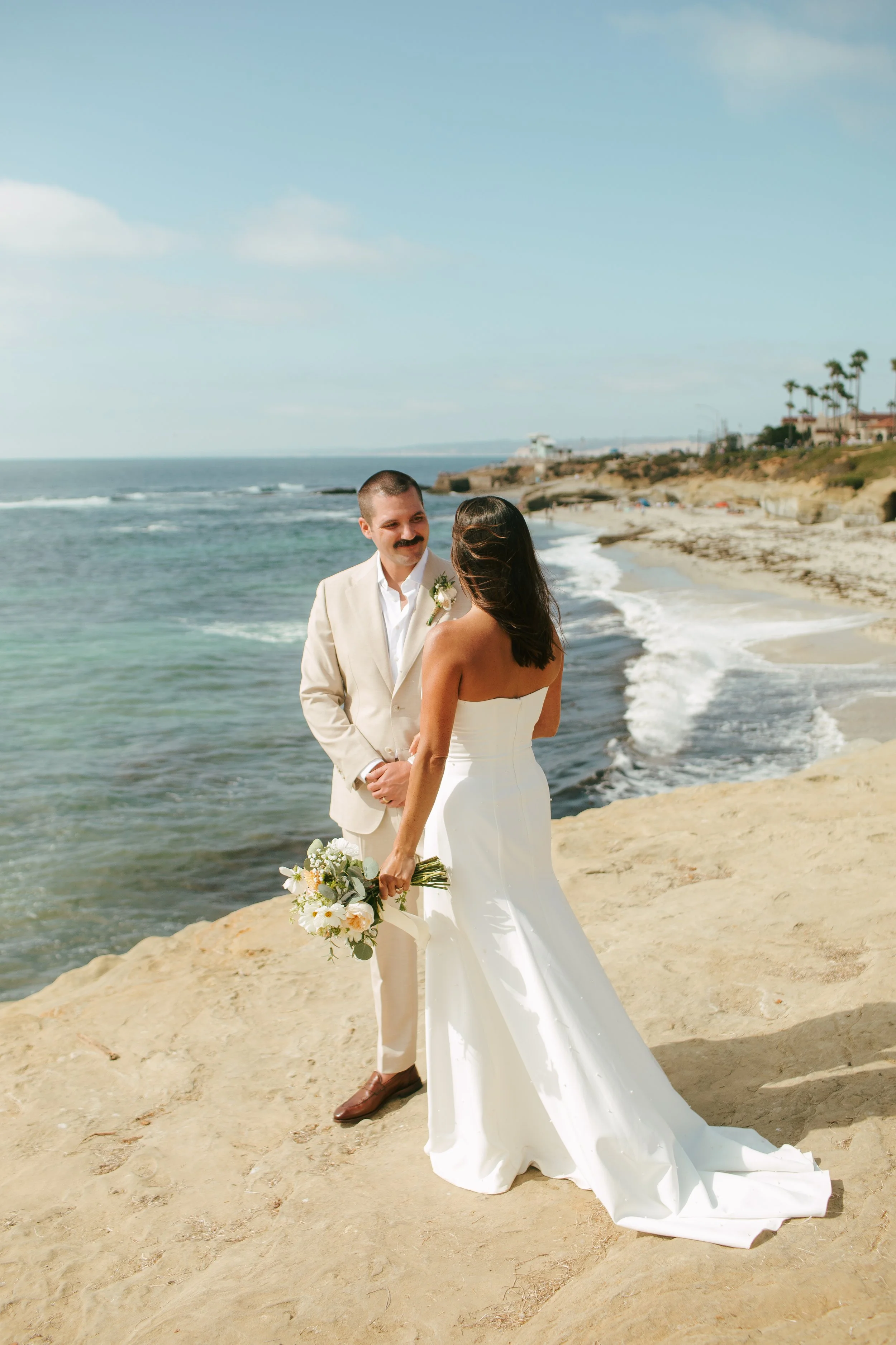A bride and groom stand on a sandy beach by the ocean, facing each other and holding hands, during their wedding ceremony, with houses and palm trees in the background.