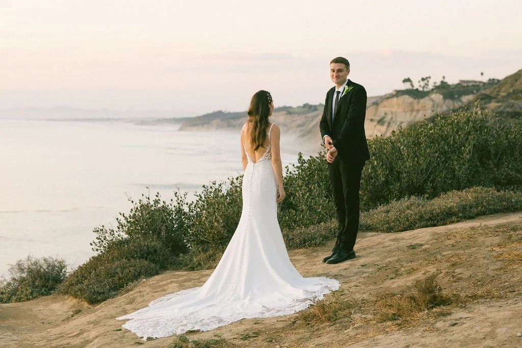 A bride and groom standing on a cliffside overlooking the ocean during sunset.