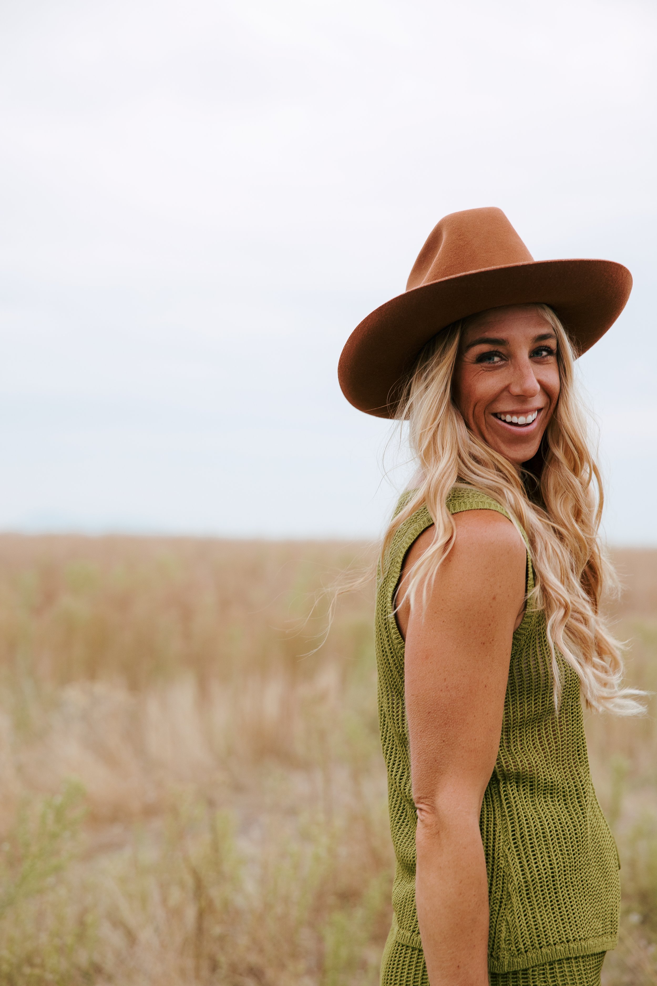 A young woman with long blonde hair wearing a brown wide-brimmed hat and a green sleeveless dress, smiling in an open field.