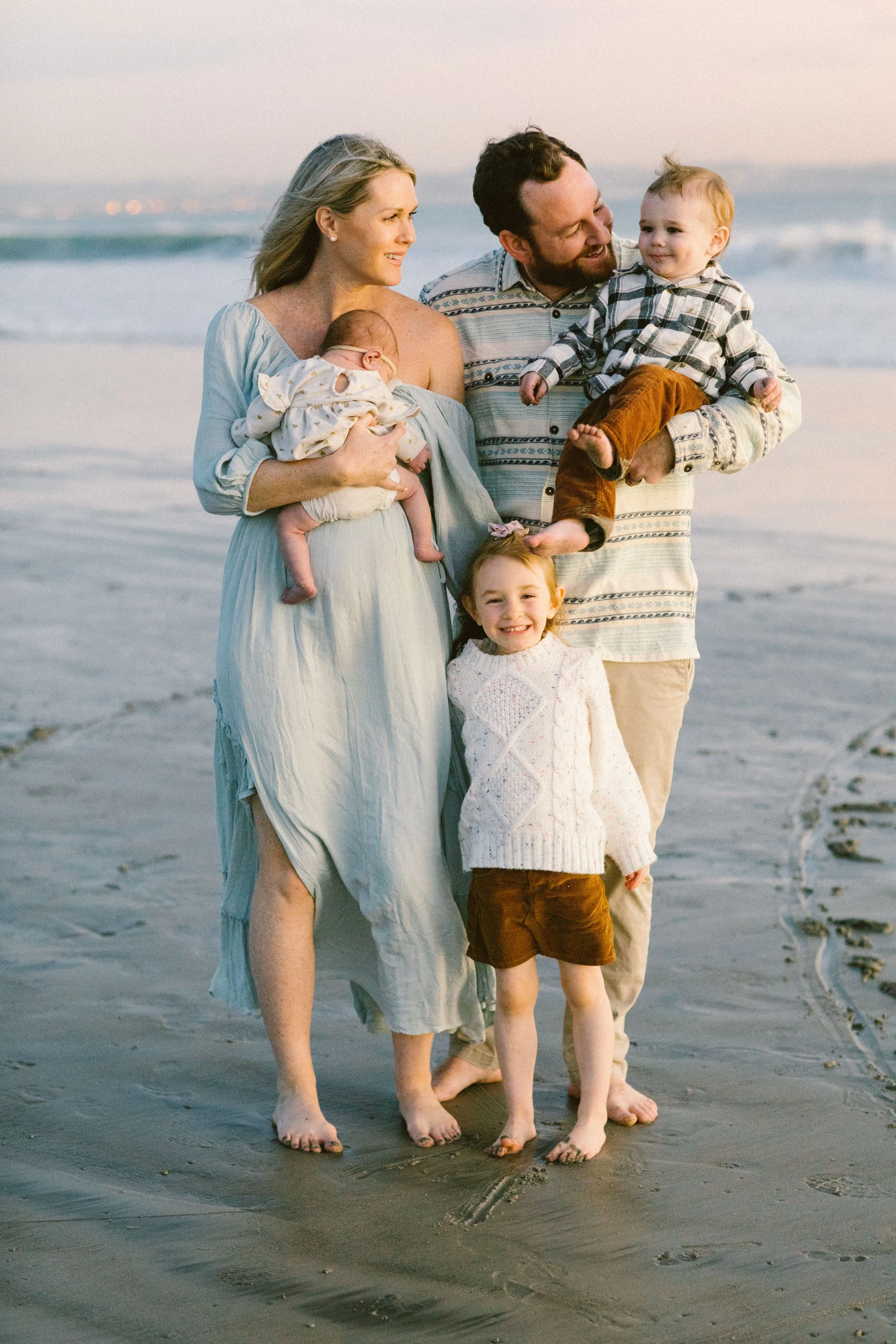Family of five on the beach, including a woman holding a baby, a man holding a young boy, and a girl standing in front, smiling as they walk barefoot on the sand at sunset.