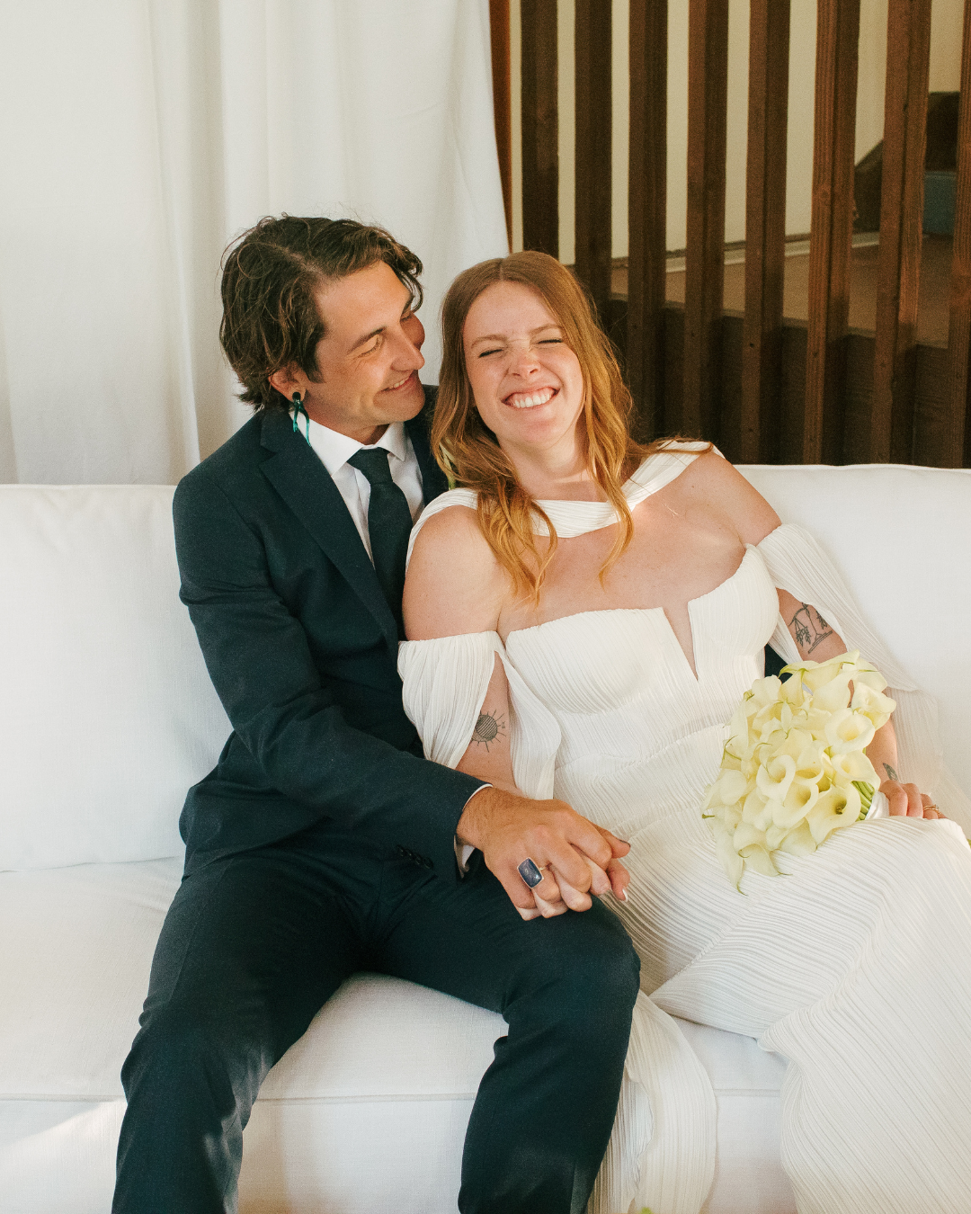 A couple sitting on a white sofa, smiling and holding hands, with a woman in a white dress holding a bouquet of white flowers, possibly at a wedding or celebration.