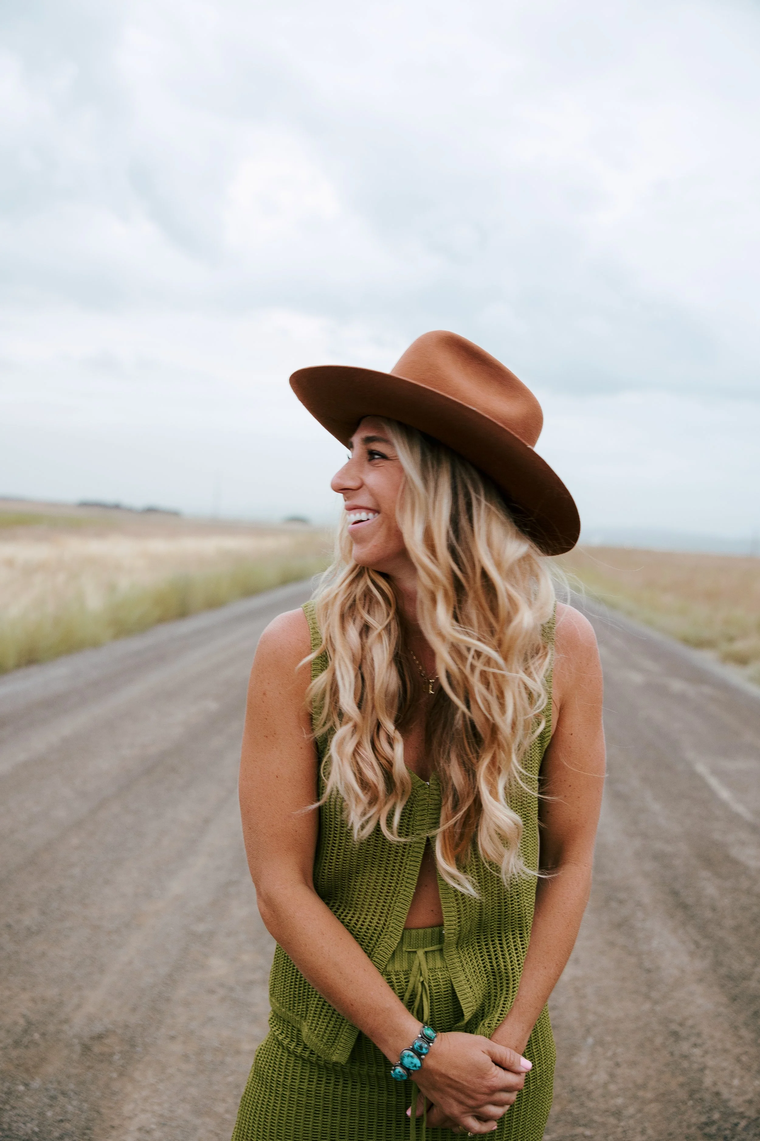 A woman with long curly blonde hair wearing a wide-brimmed brown hat and a green sleeveless dress, smiling and standing outdoors on a dirt road with open fields and a cloudy sky in the background.