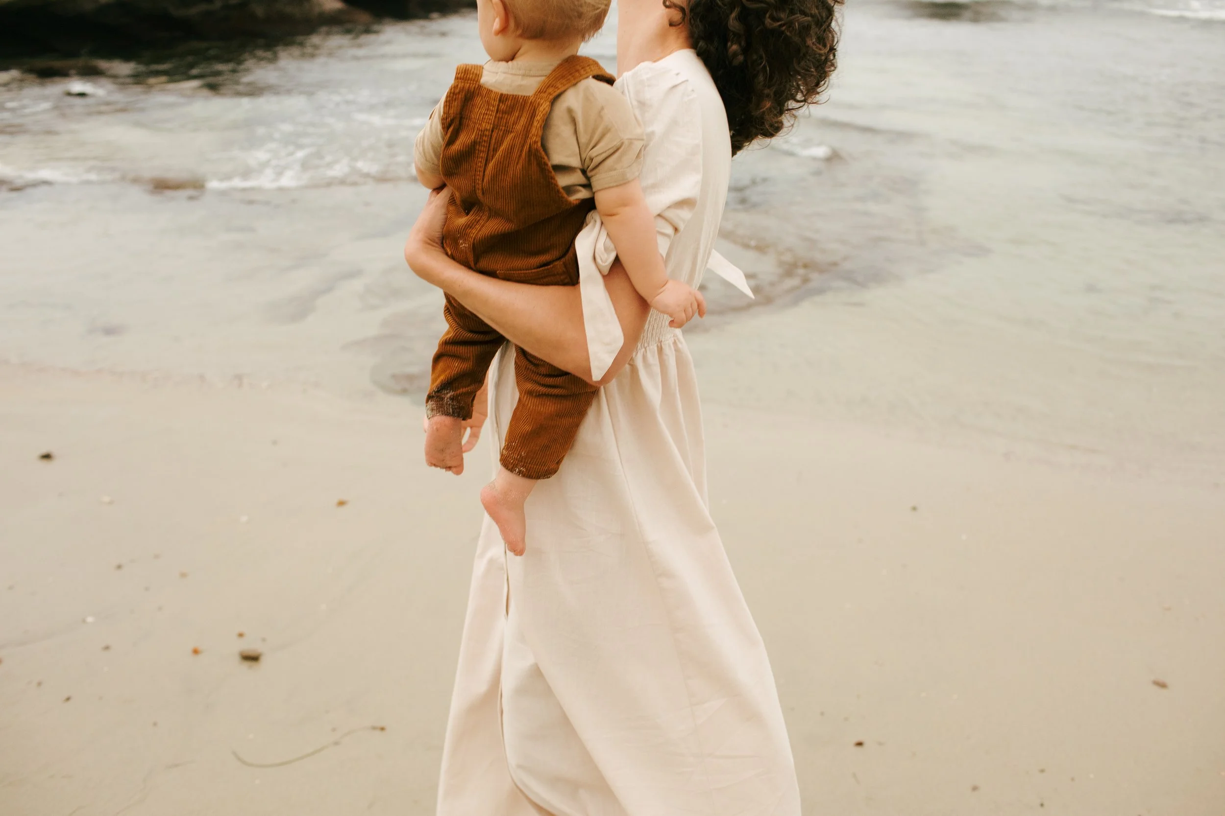 A woman holding a young child at the beach, both legs of the woman visible, wearing light-colored clothing, with shallow water and sand in the background.