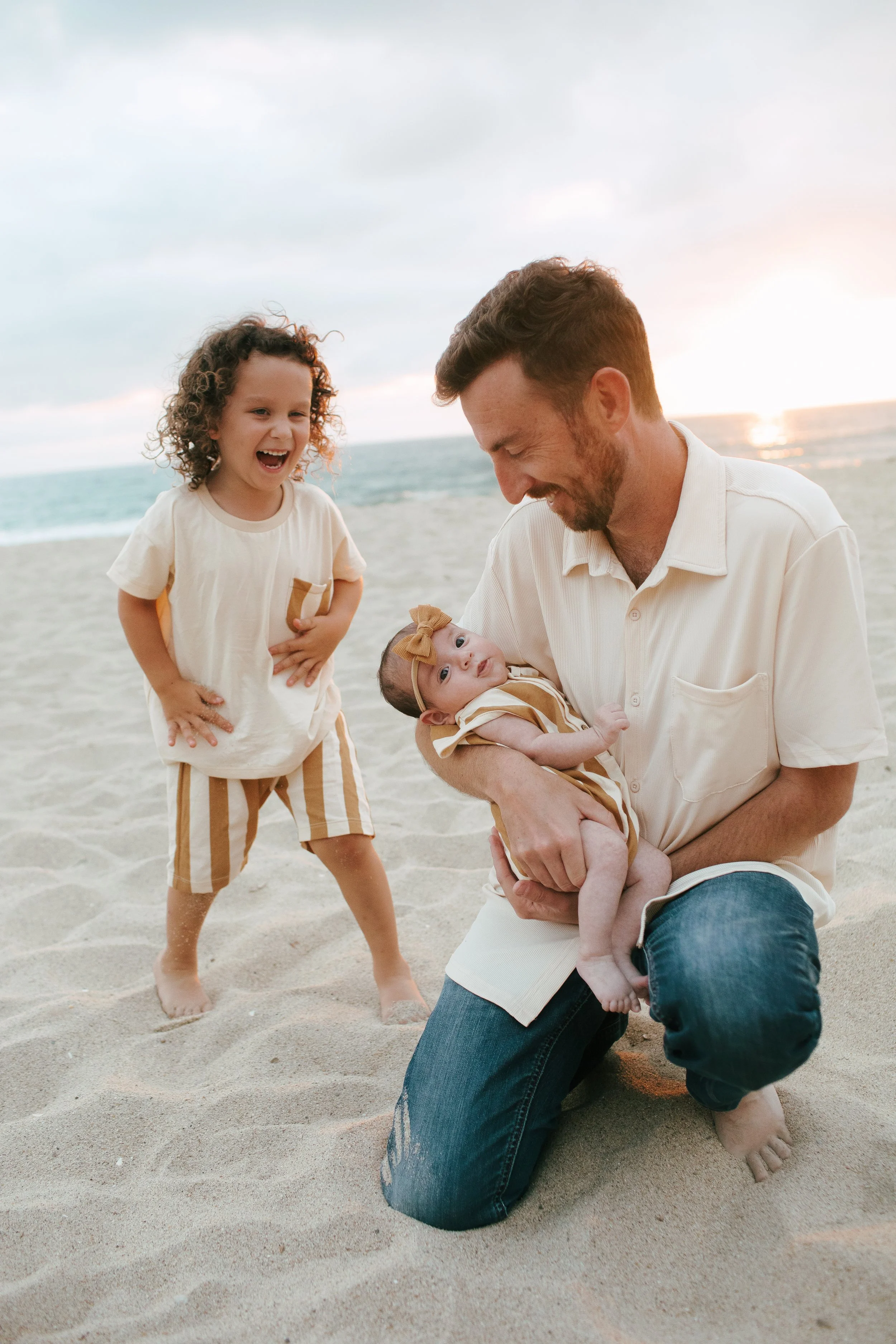 A man kneeling on the sand at the beach holding a baby girl, with an older girl smiling and laughing nearby, all enjoying a sunset.
