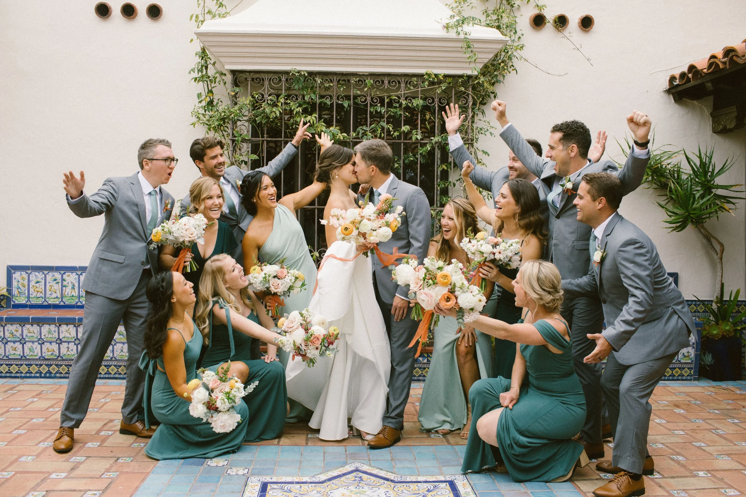 A wedding celebration with a bride and groom kissing surrounded by friends and family, all happily celebrating outdoors against a white wall with tiled steps and green plants.